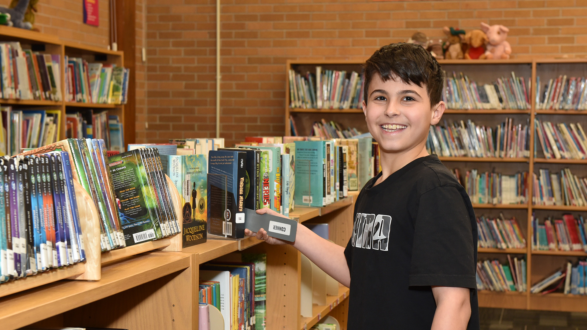 Child selecting a book from a wooden shelf in a school library filled with colorful books and brick walls.