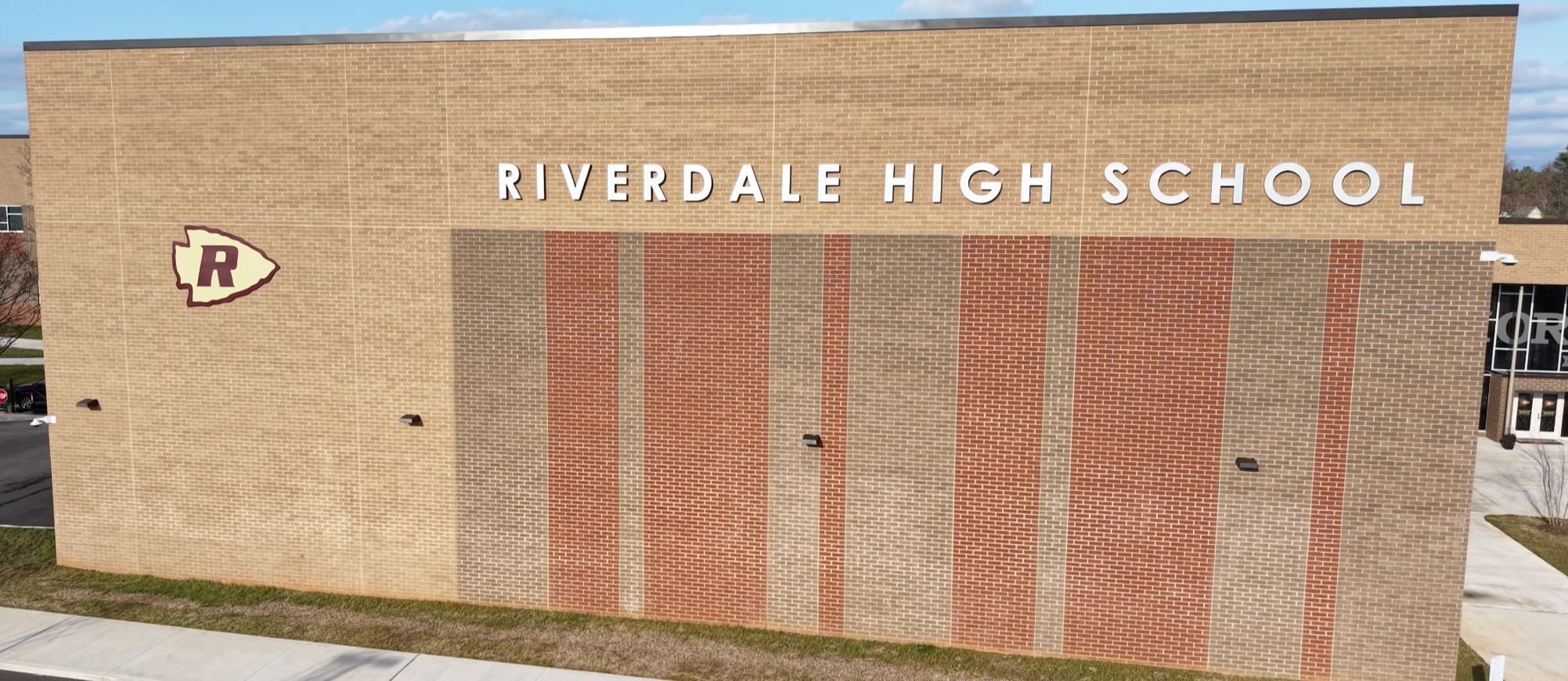 A high school building with a large "RIVERDALE HIGH SCHOOL" sign on its side. The school is beige with orange and brown stripes.