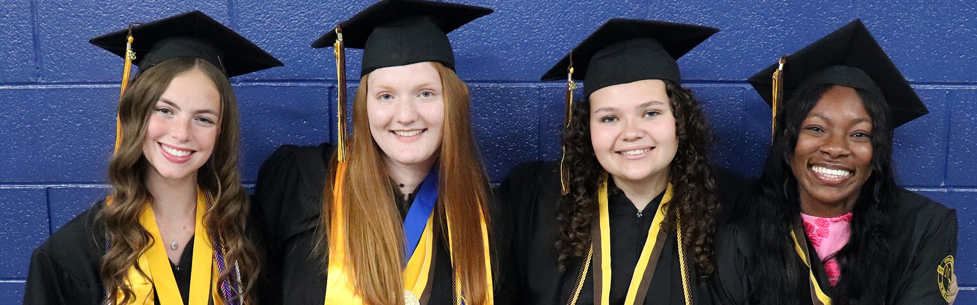 Four NFHS graduates sit next to each other smiling in their cap and gowns