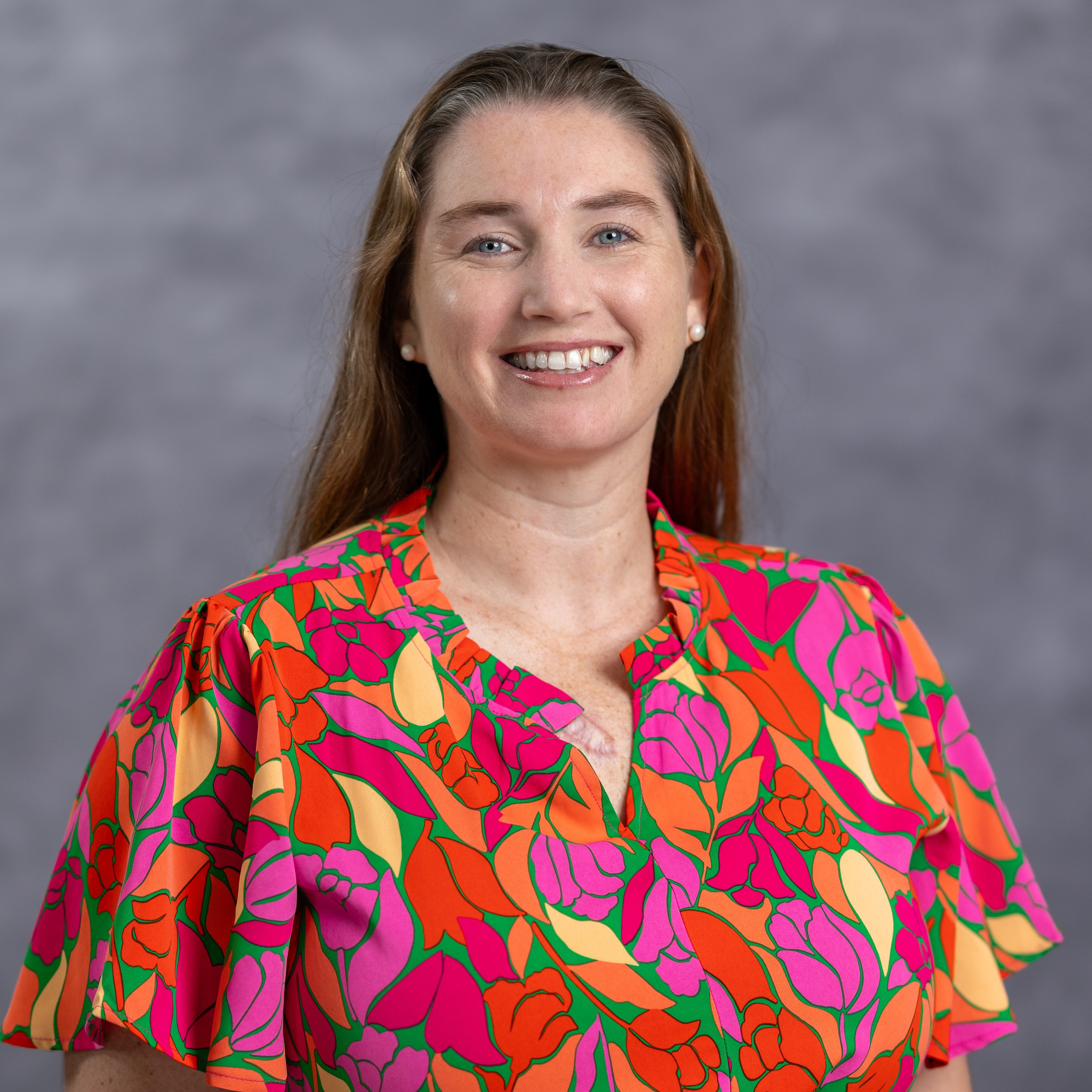 Woman with long brown hair smiling, wearing a short-sleeved floral shirt, and standing against a gray background.