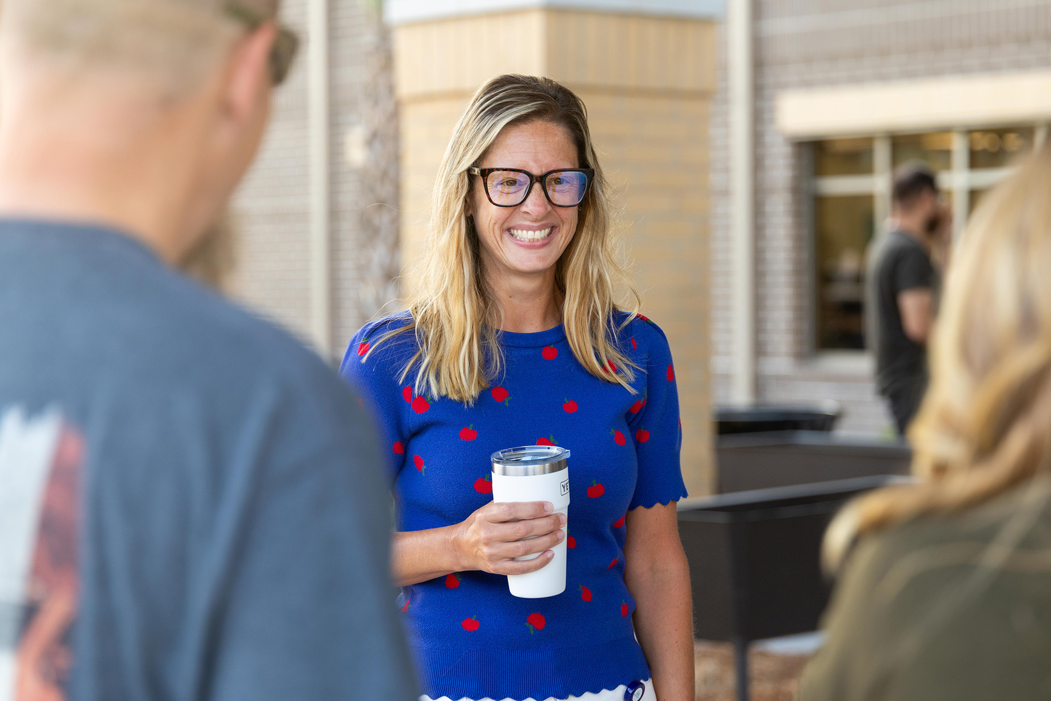 A woman in a blue polka-dot dress holds a cup in a courtyard with two others.