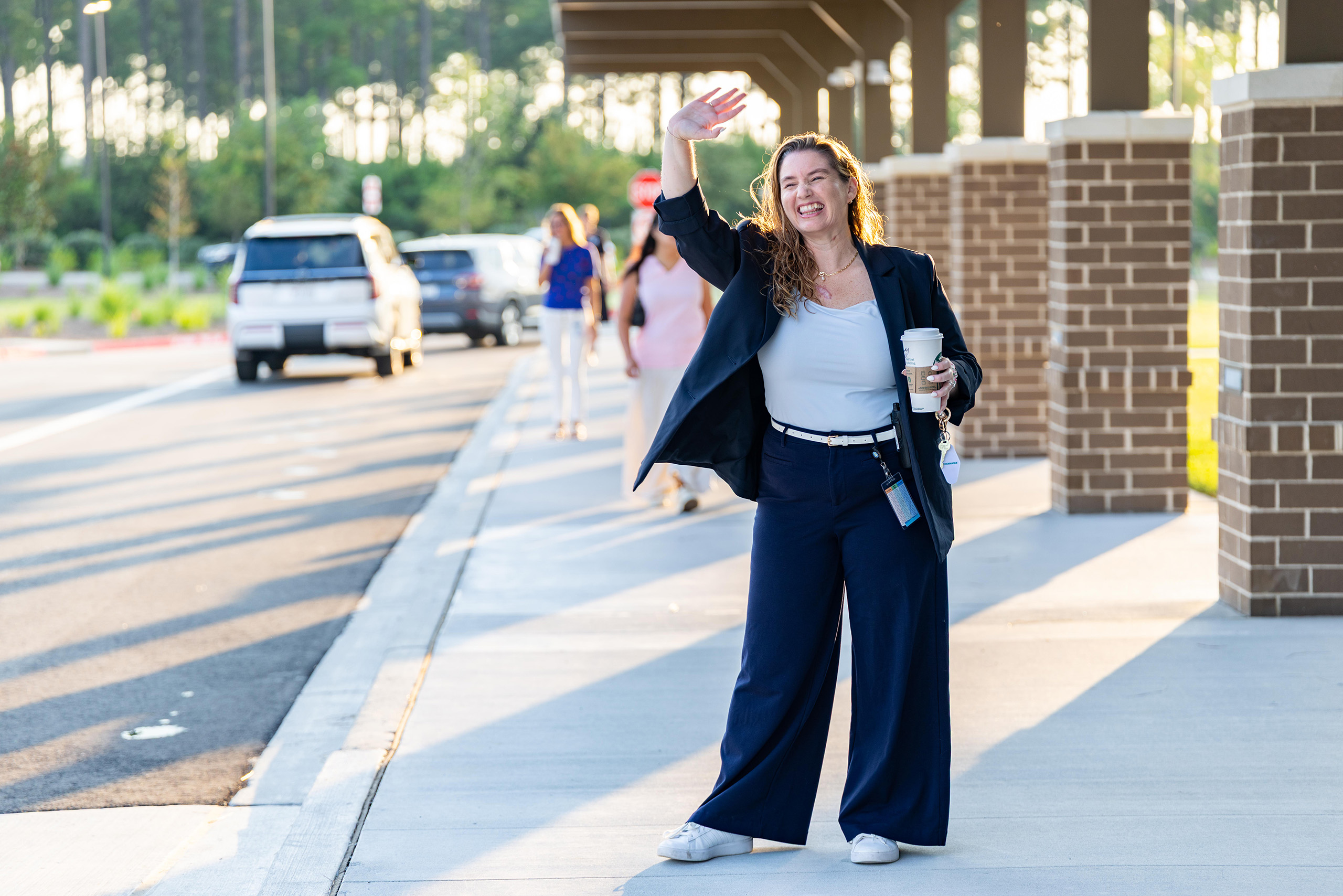 A woman waves while standing on a sidewalk. She wears a white top, blue pants, and a jacket.