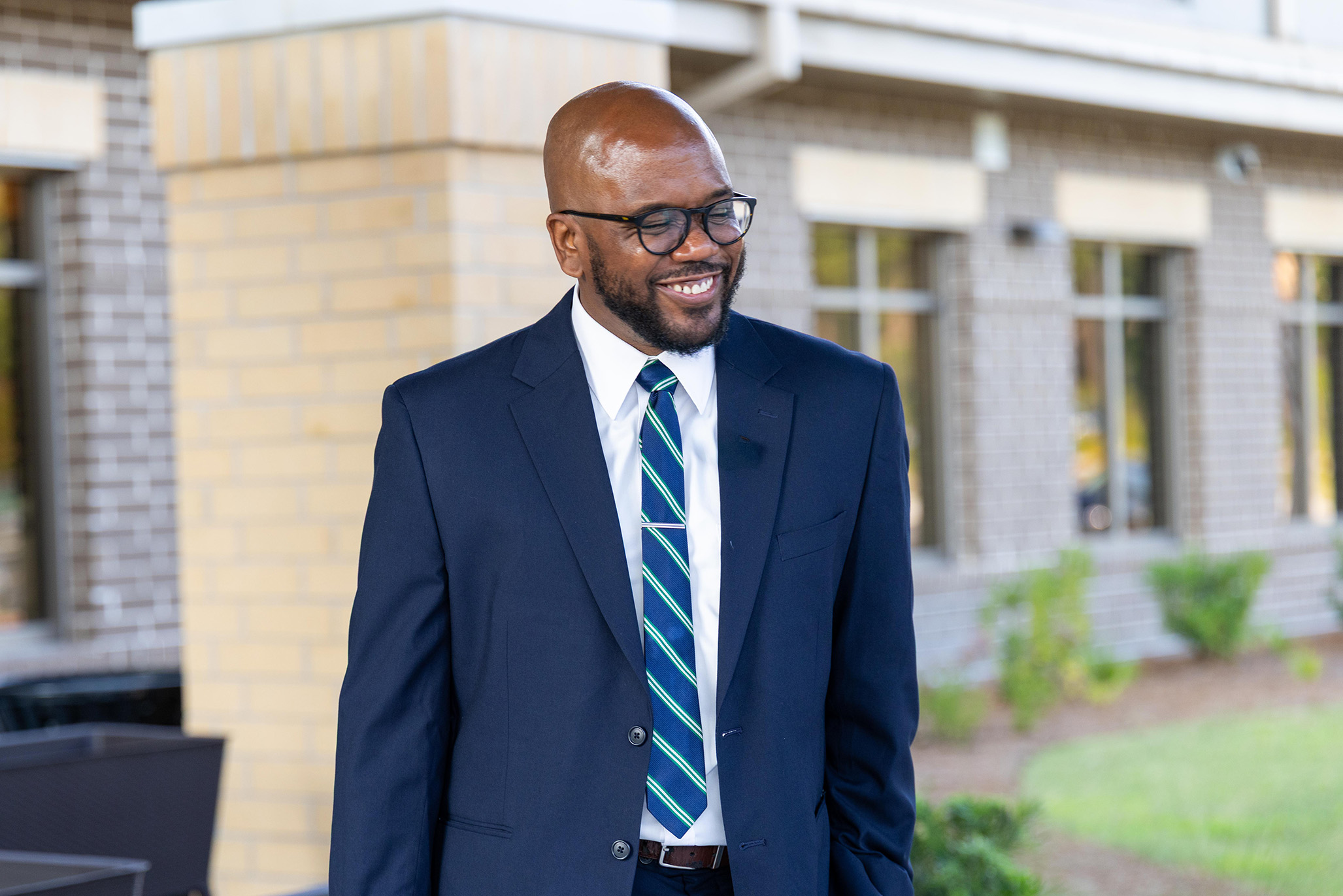 Man in a navy suit with a striped tie and glasses smiles in front of a building.