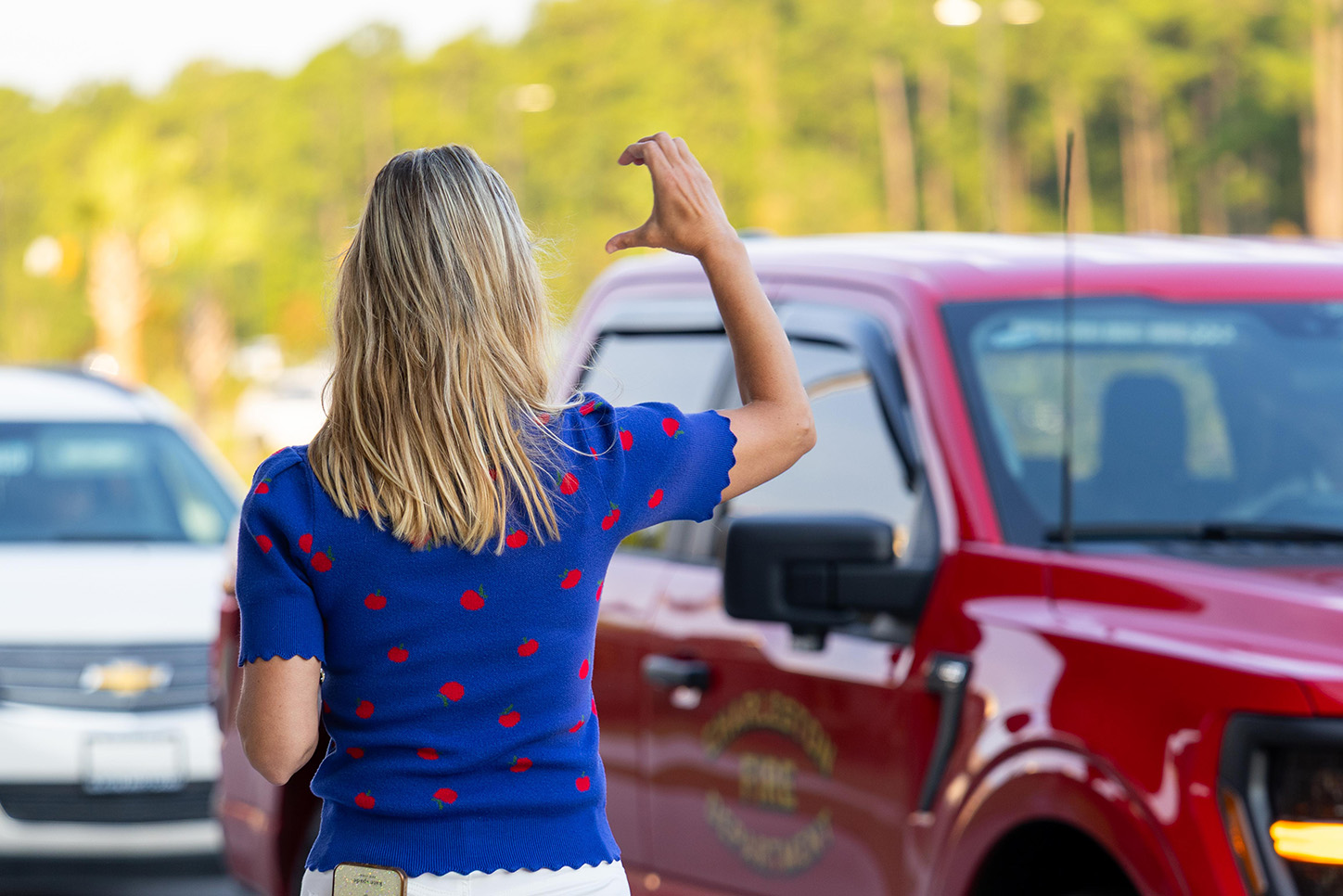 A woman with blonde hair waves from the passenger side of a red truck. Trees in the background.