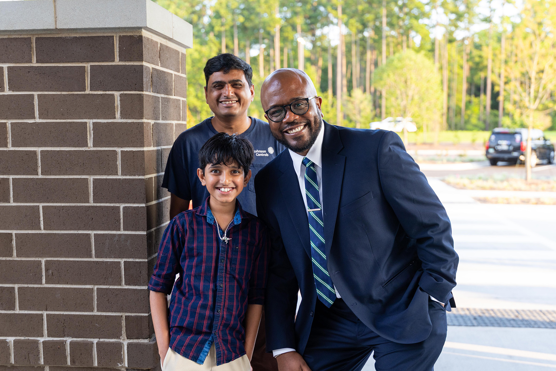 Three people in business attire stand by a brick column. Behind them, a road with greenery and parked cars is visible.