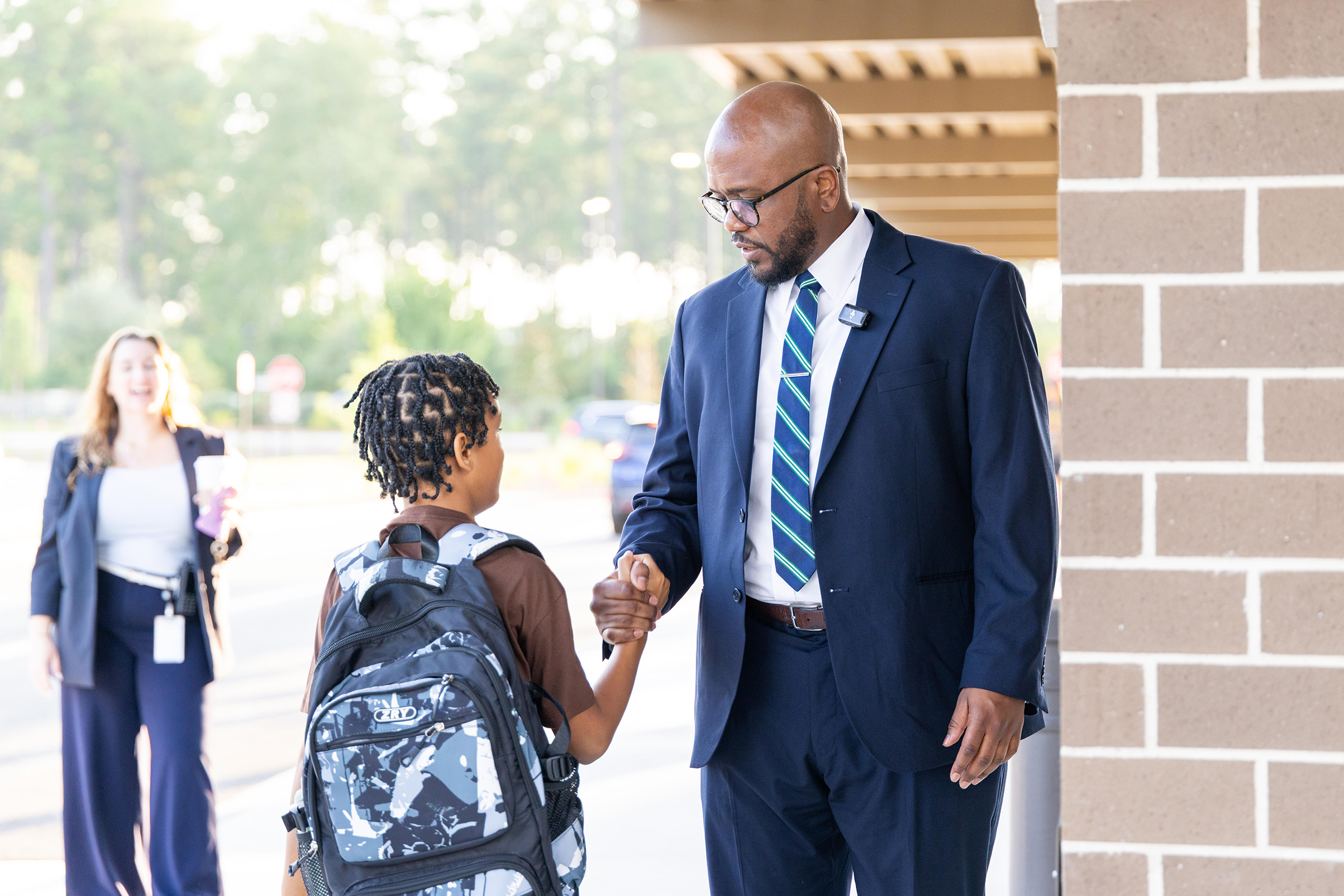 A man in a suit shakes hands with a child carrying a backpack. A woman walks behind them. Trees are in the background.