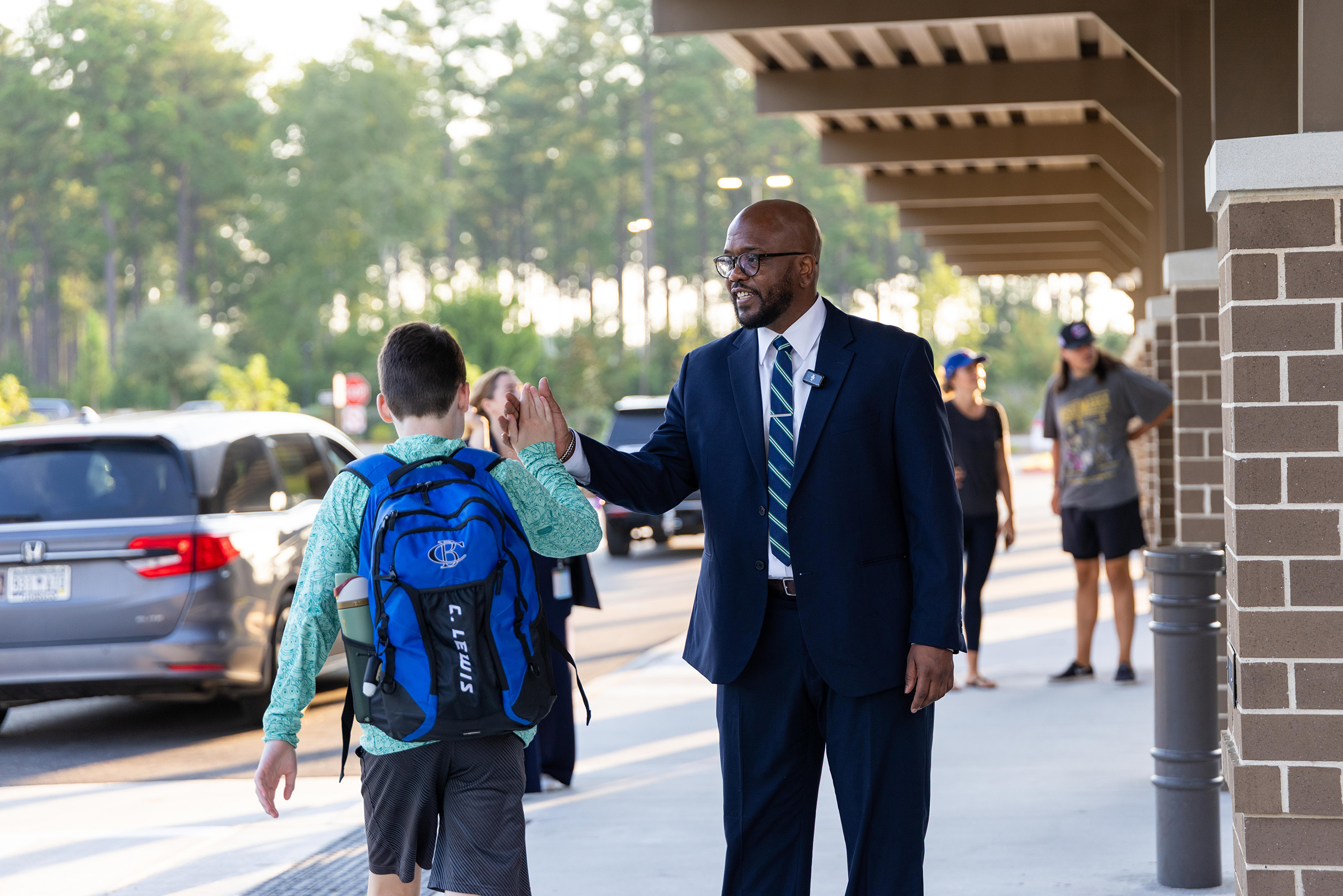 Adult man in a suit and tie shakes hands with a boy in a backpack. People walk in the background.