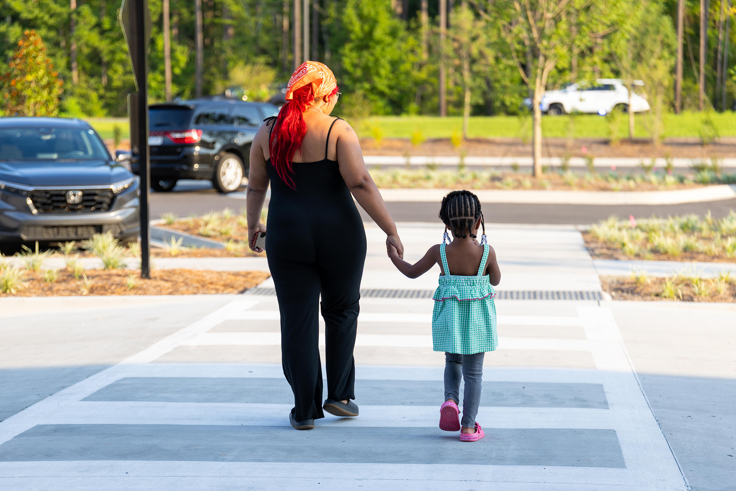 An adult and a child walk hand-in-hand on a pedestrian crossing, with cars parked in a parking lot.