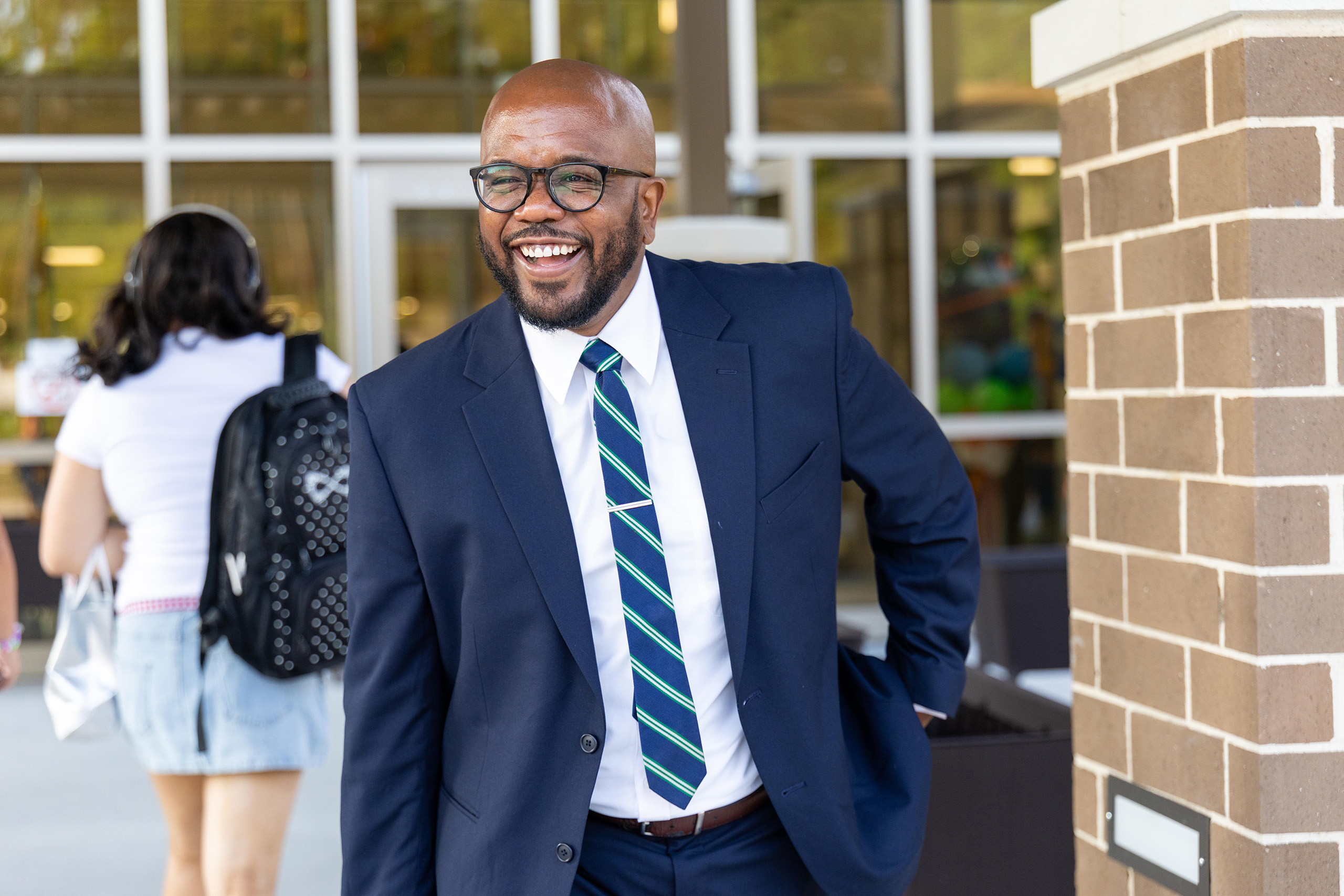 A man in a dark suit, glasses, and a striped tie stands with one hand in his pocket. Behind him, a woman walks with a backpack.
