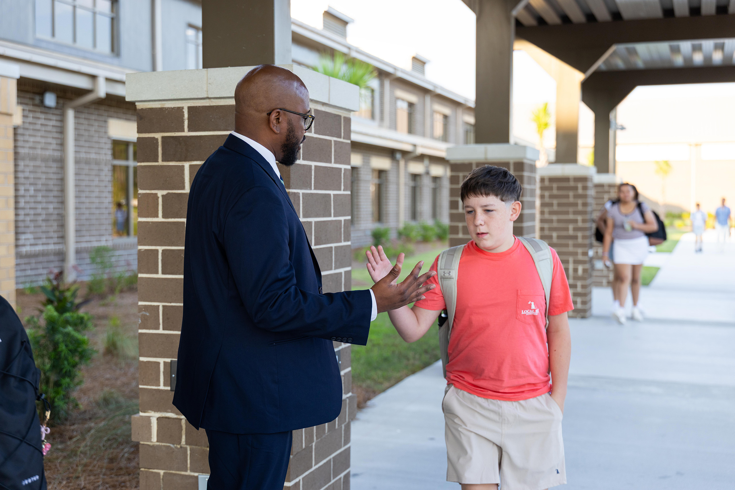 Man in a suit talks to a boy wearing a backpack at a school entrance.