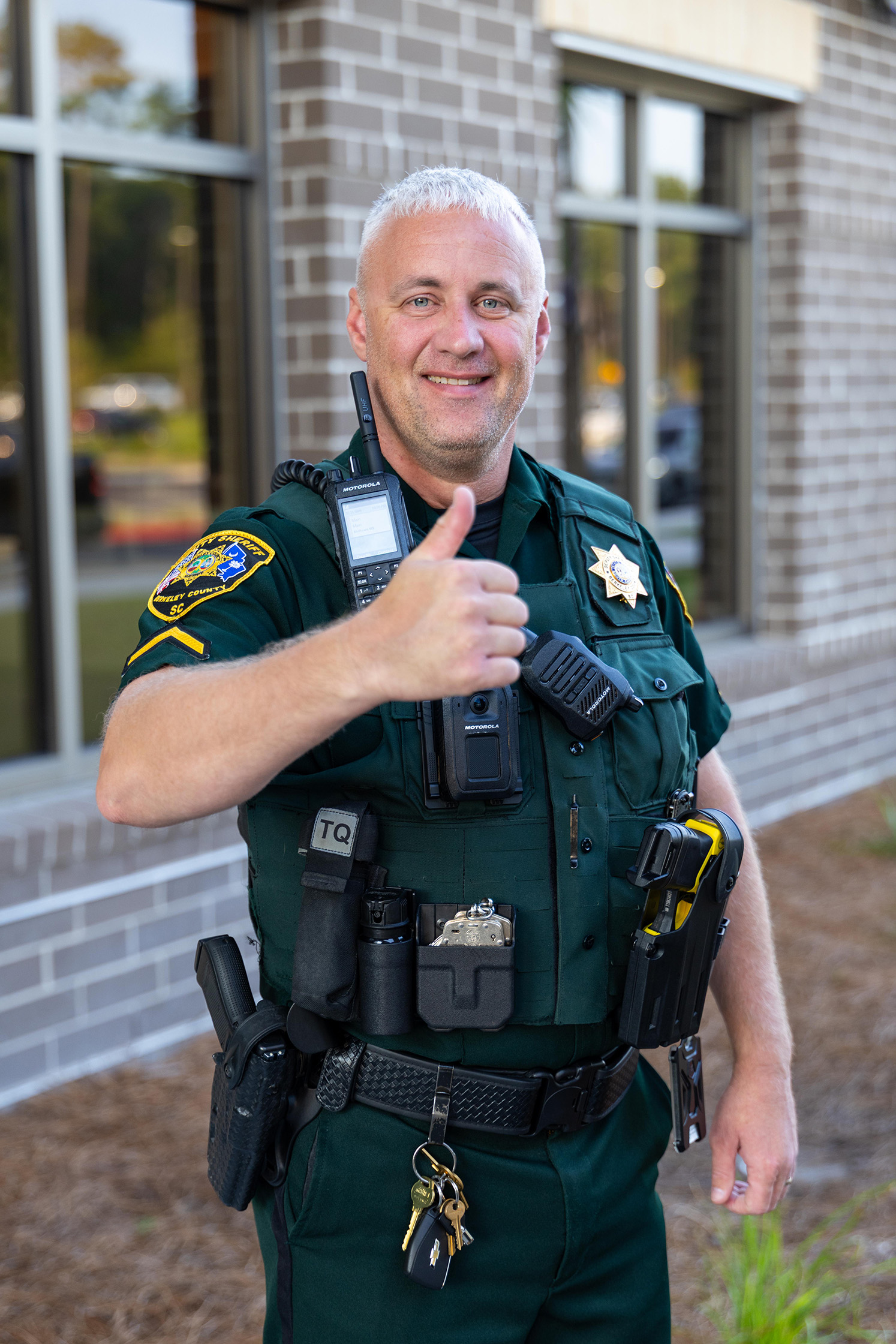 A police officer, in uniform, smiling and giving a thumbs up. He stands in front of a building.