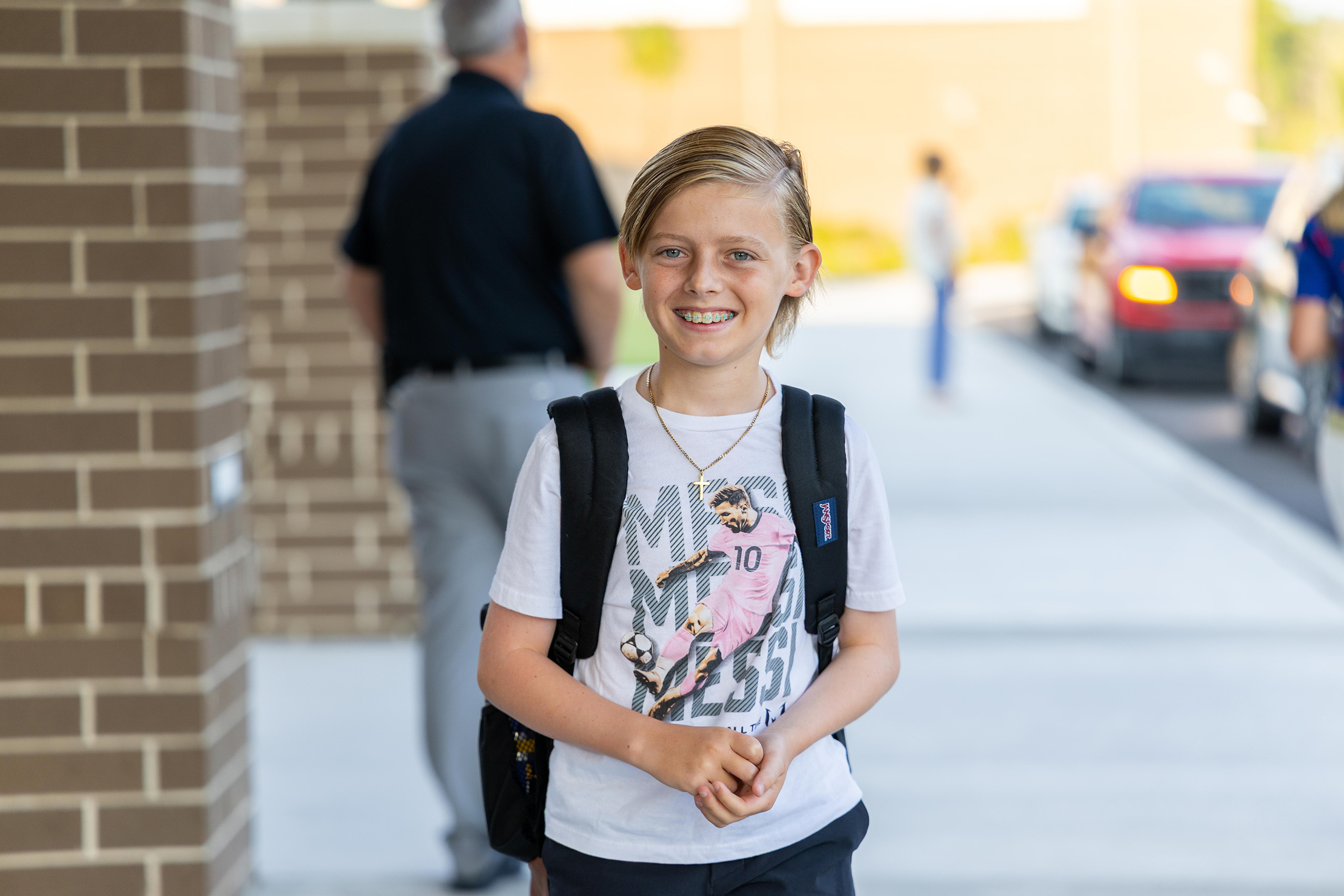 Child in white T-shirt with graphic print, black backpack, and smiling face standing on sidewalk.