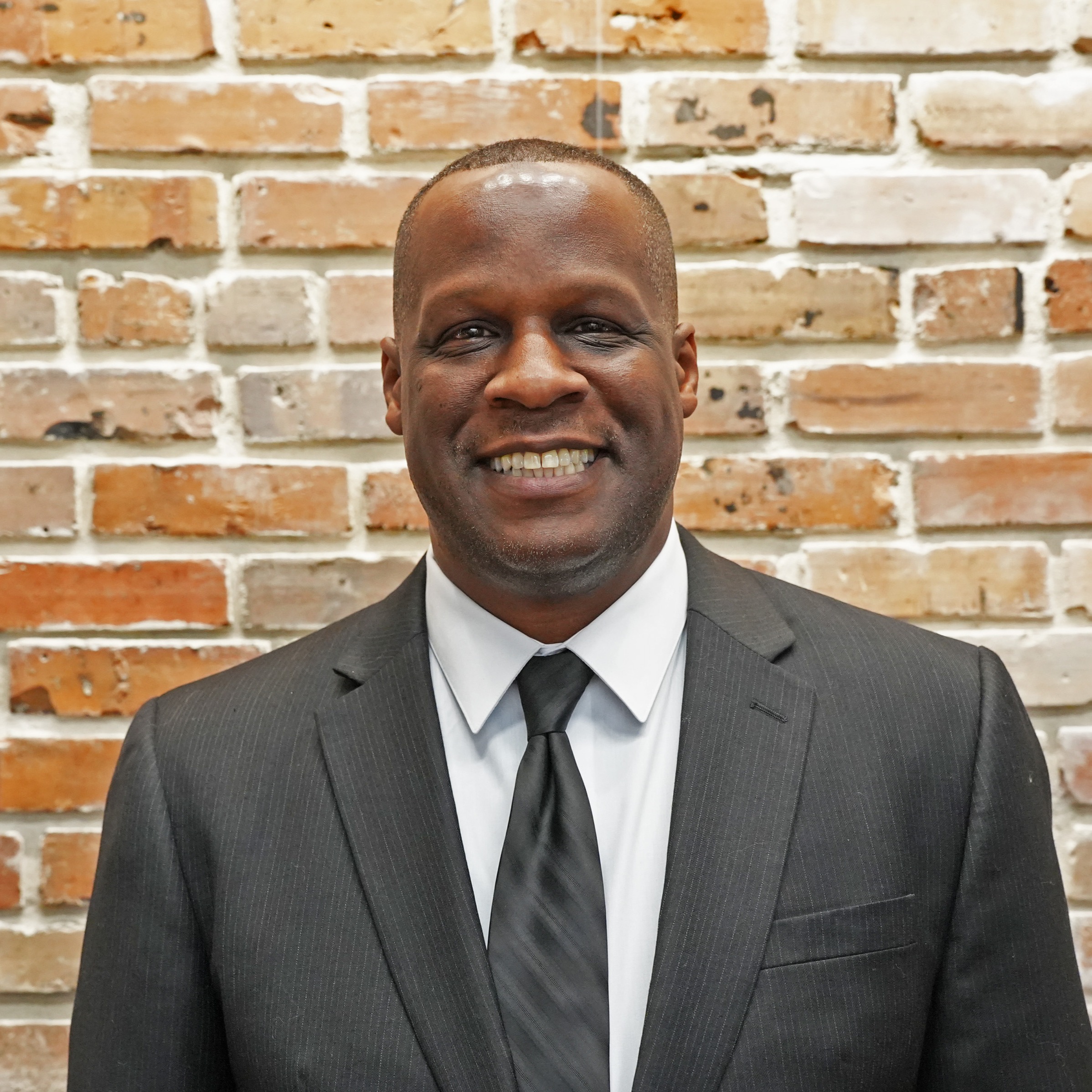 A man in a suit and tie stands in front of a brick wall, smiling.