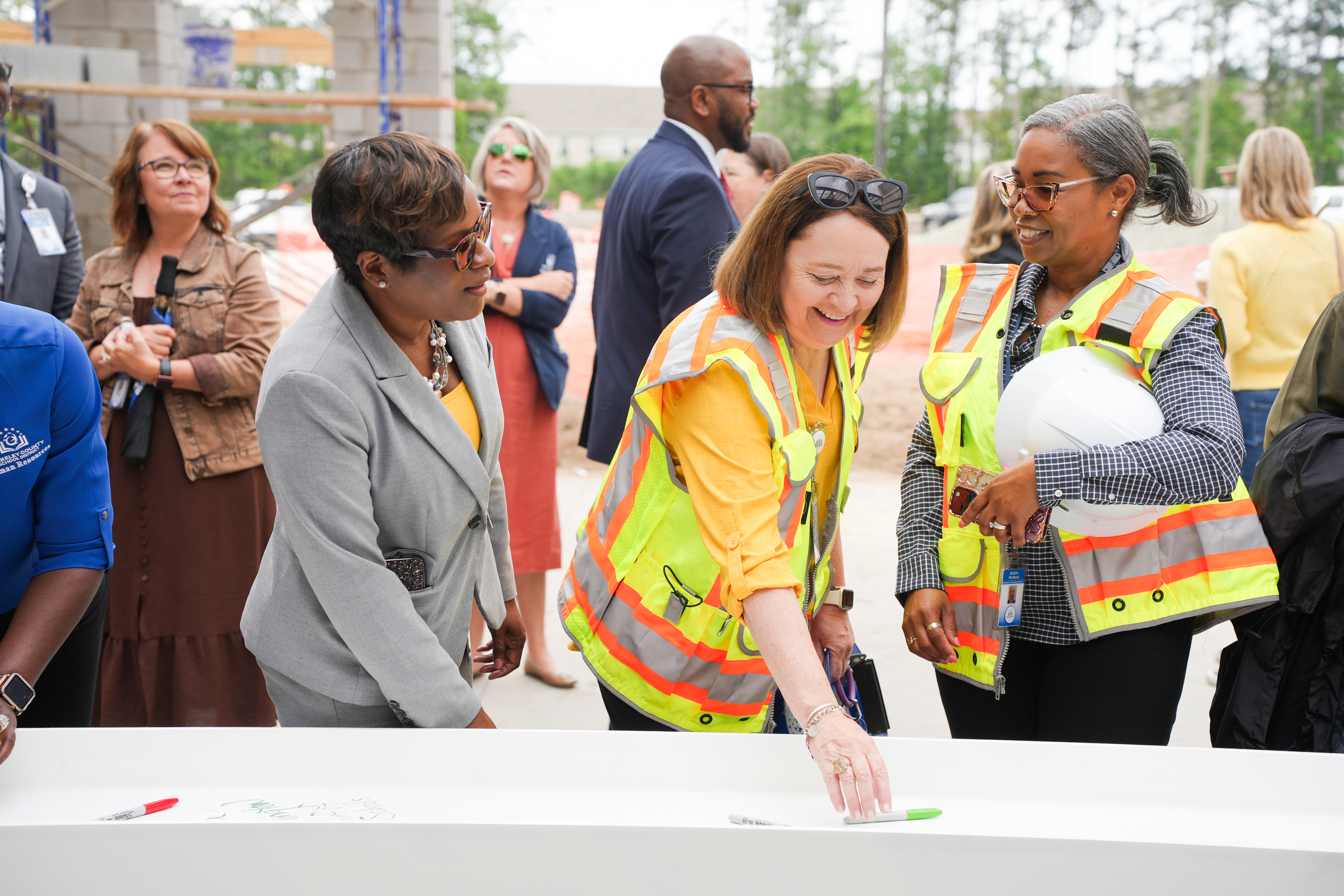 A group of adults with reflective vests and hats are gathered around a table at a construction site.