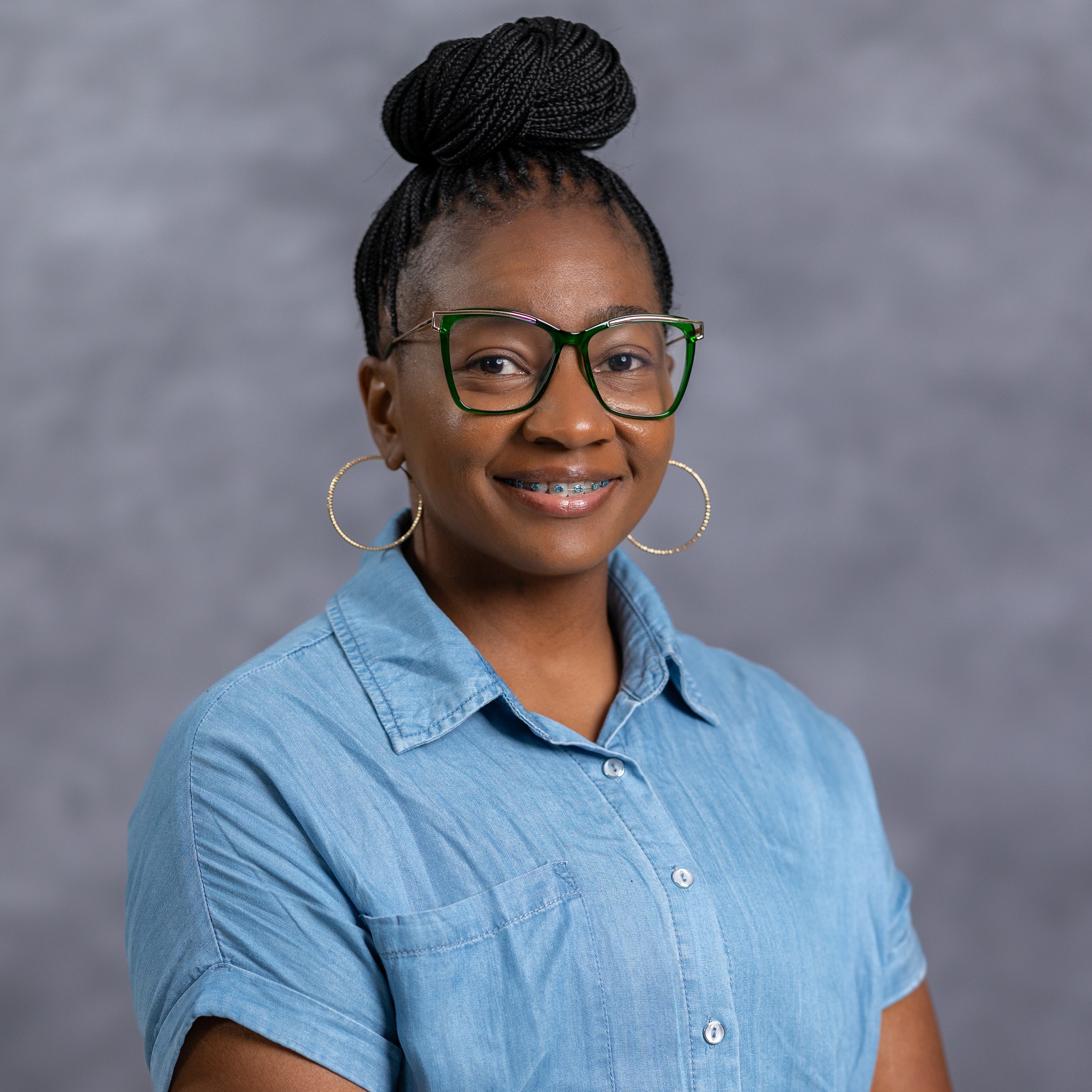 A smiling woman with glasses and large hoop earrings poses for a photo against a gray background.