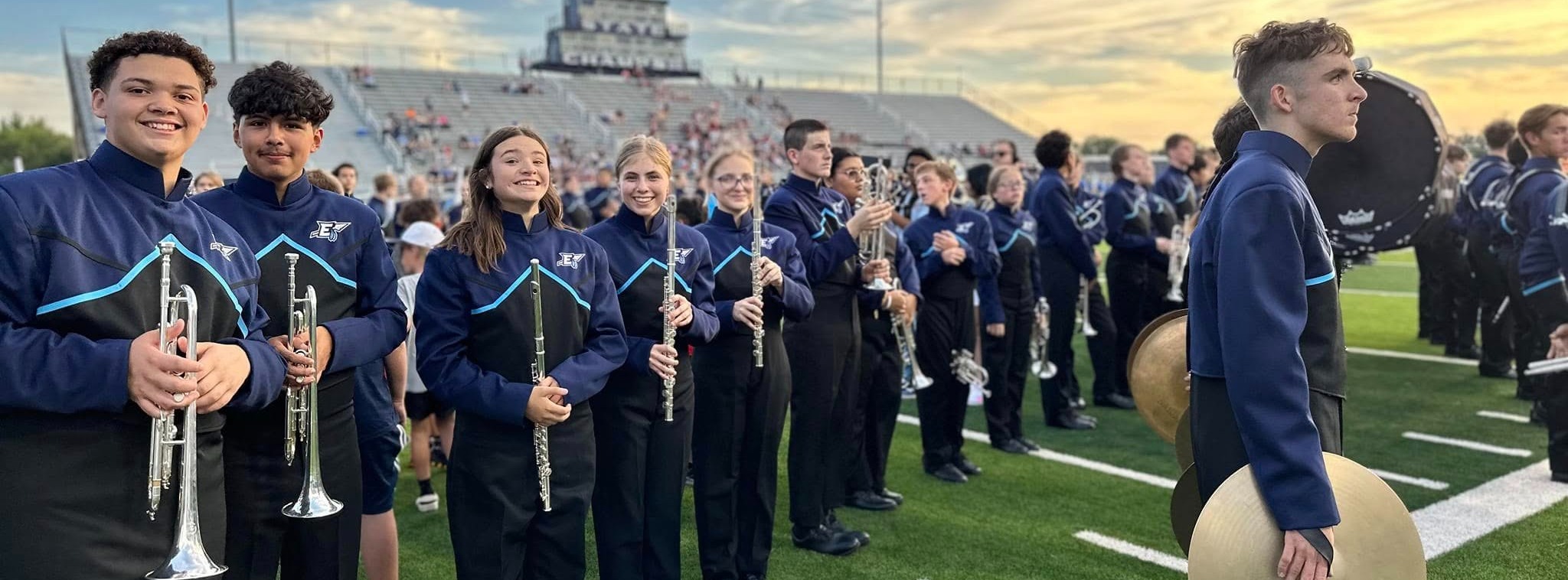 Enid High School marching band students in matching blue uniforms stand in rows on the football field with instruments 