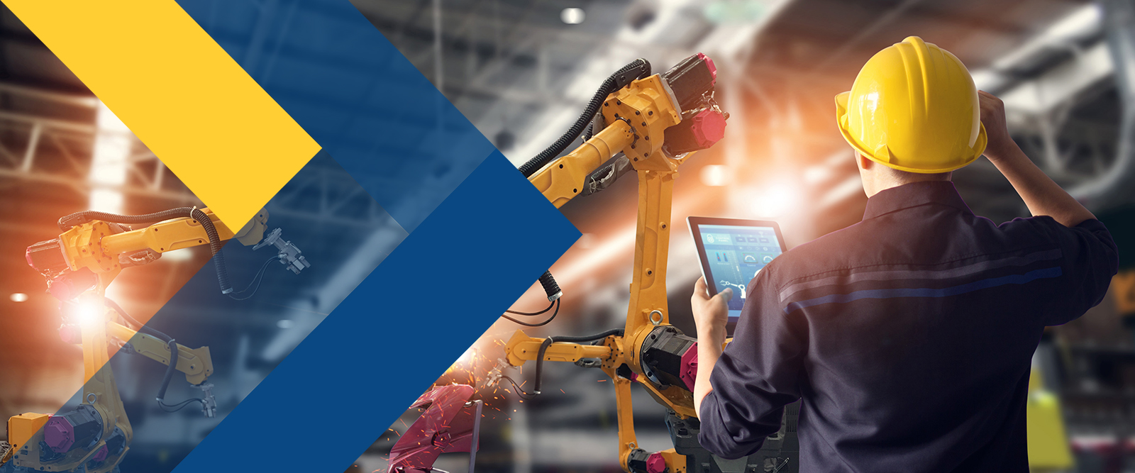A person in a yellow hard hat using a tablet stands in a factory with robotic arms.