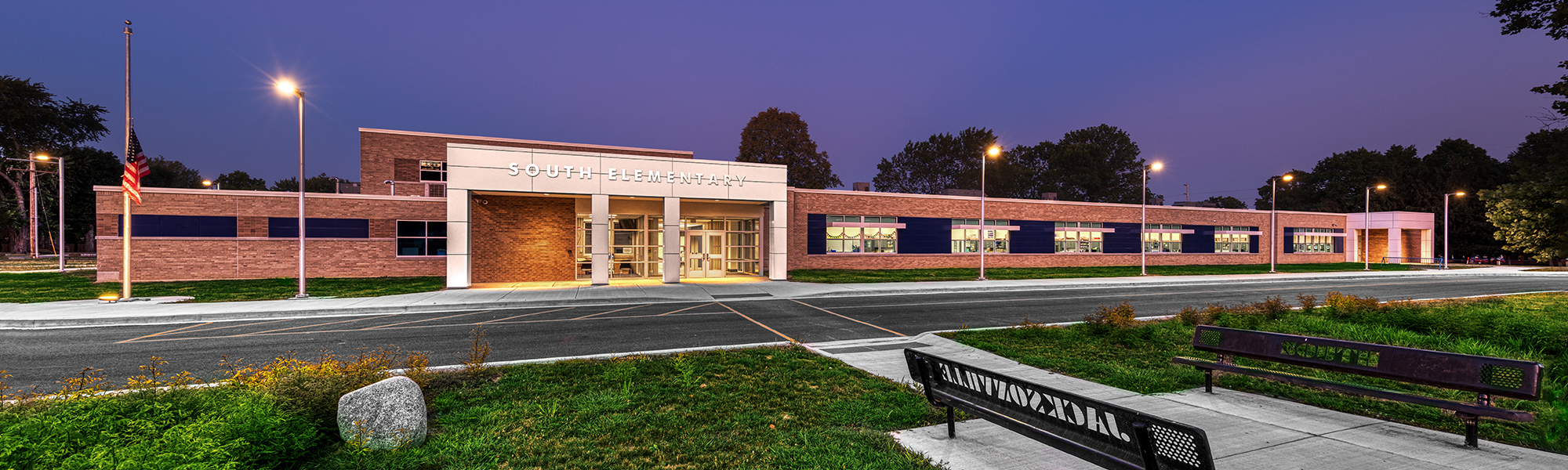A wide view of South Elementary shows the modern brick school building at dusk, with a brightly lit main entrance, landscaped grounds, an American flag, and benches in the foreground overlooking the driveway.