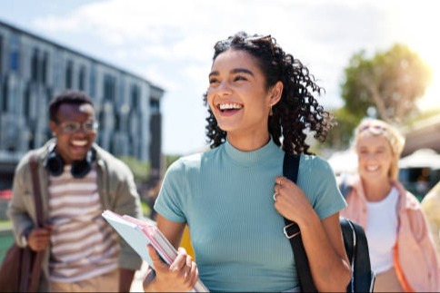 Three smiling high school students walk together across a school campus on a sunny day. The student in front carries notebooks and wears a backpack, while the others follow behind with backpacks and headphones. Modern school buildings and trees are visible in the background, creating a welcoming, academic atmosphere.