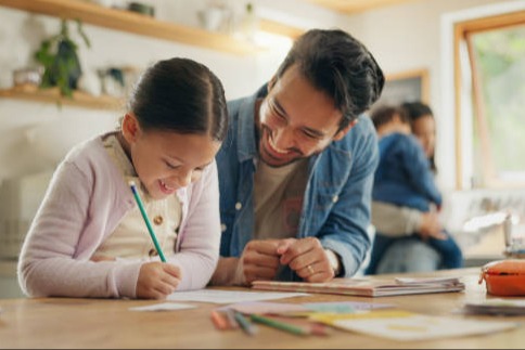 An adult helps a young student complete schoolwork at a kitchen table while the child writes with a pencil. Colored pencils and school supplies are spread across the table, and two other family members are visible in the background, creating a warm, supportive home learning environment.