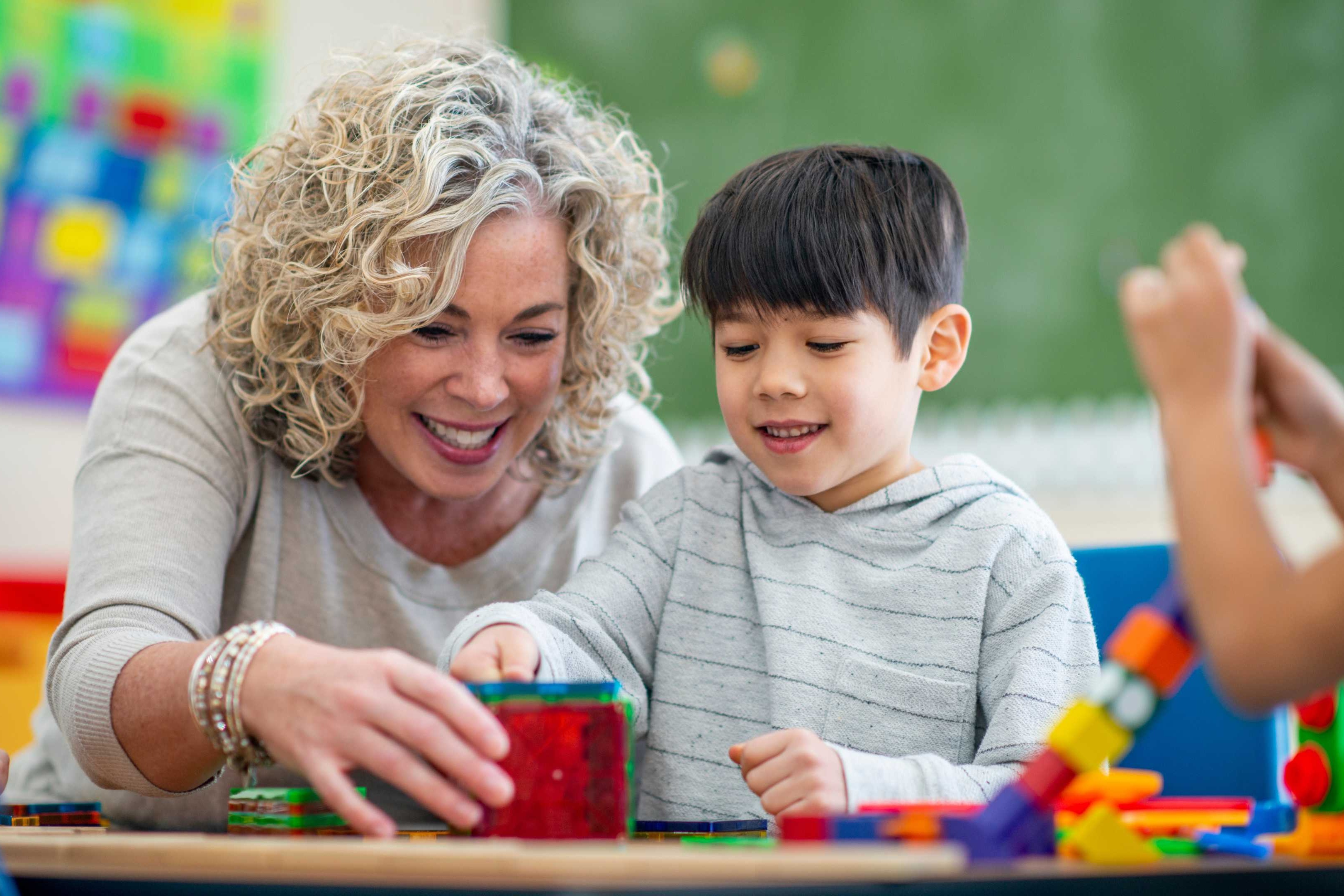 A woman smiles while sitting with a child. They are at a table with building blocks.