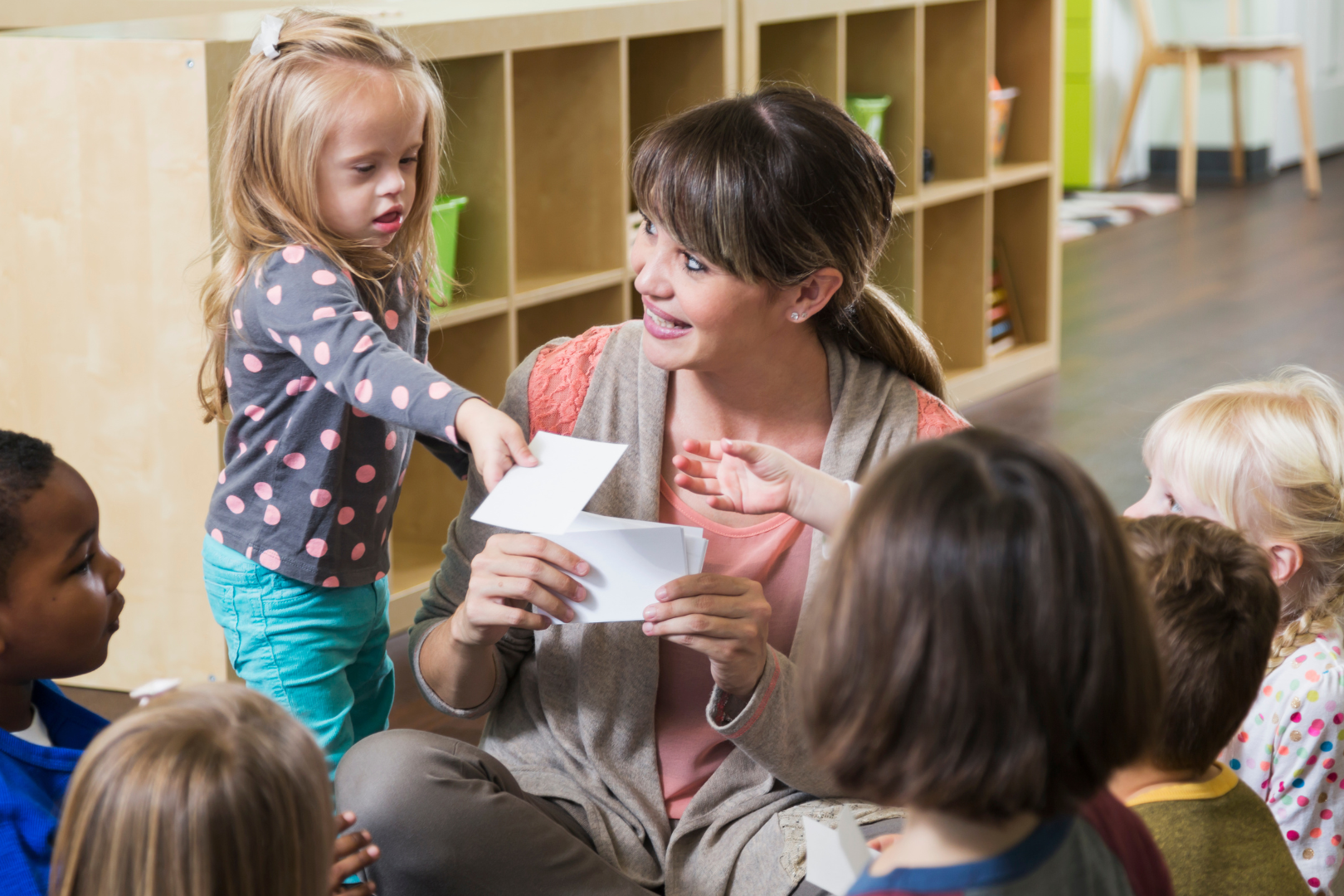 Woman in circle with children. Woman holding paper, children reach for paper. Background has shelves and chairs.