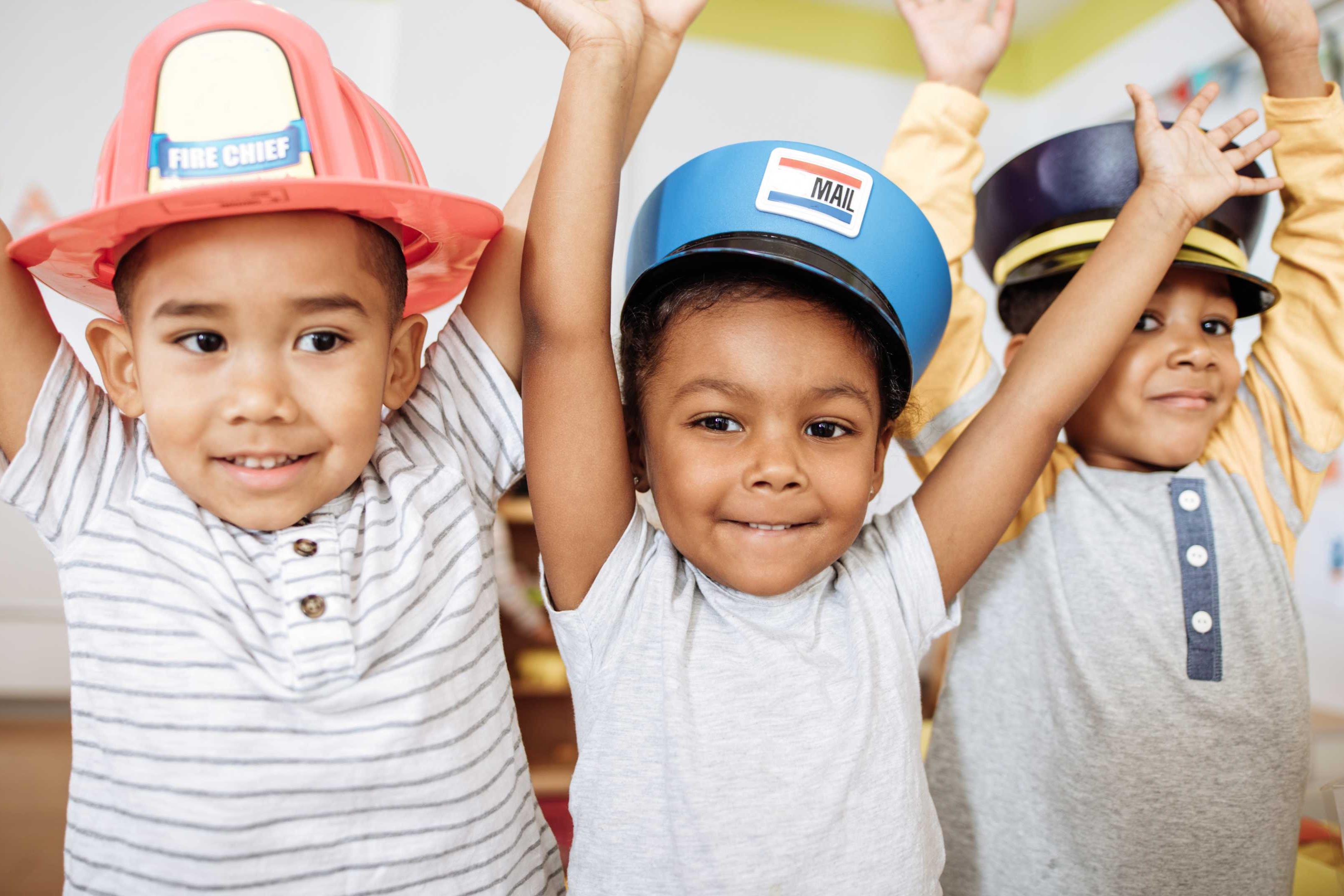 Three children in hats. One wearing a red and white fireman hat, another a blue hat, and the third a black hat.