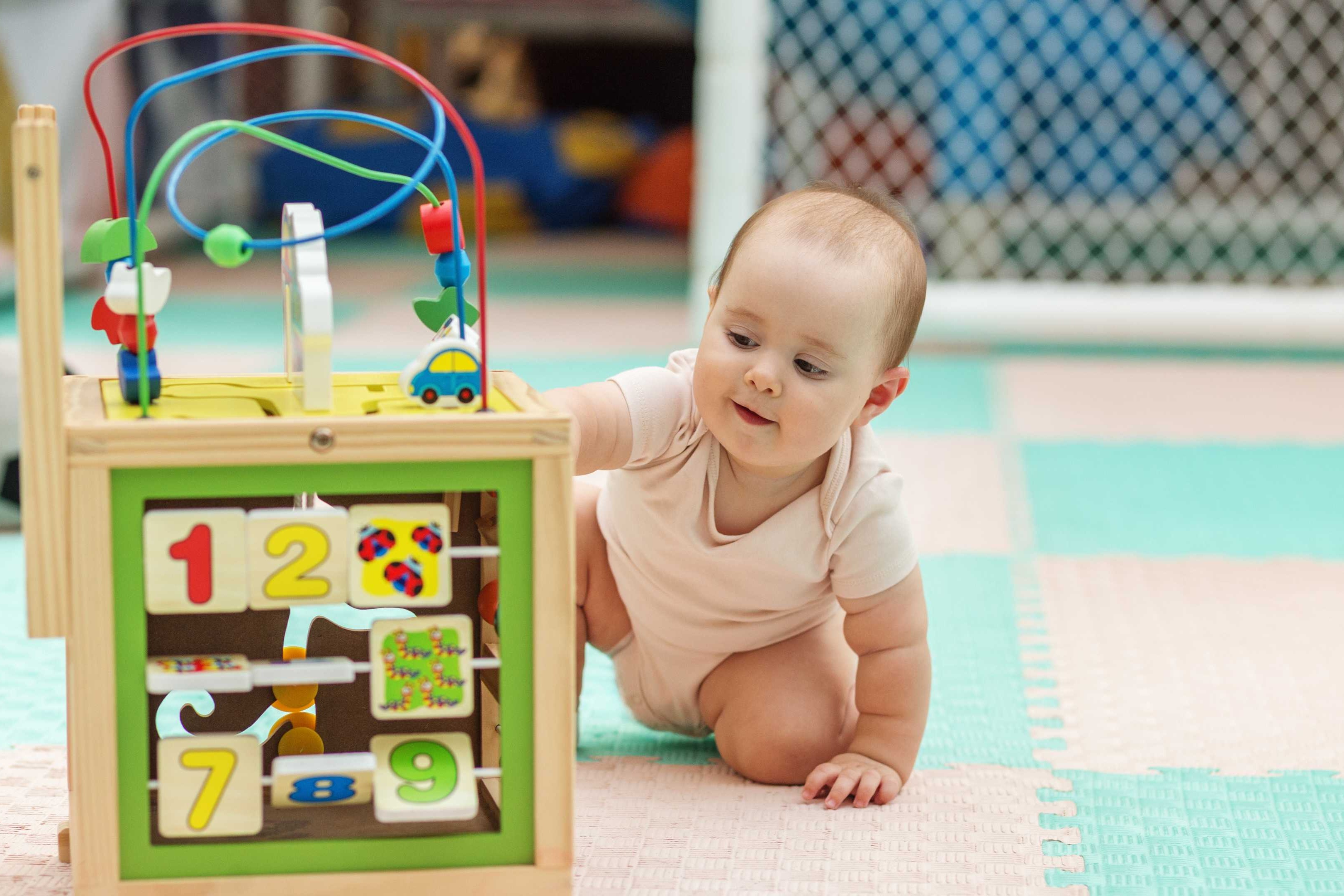 A baby plays with a colorful wooden toy with numbers, shapes, and balls on a multi-colored floor.