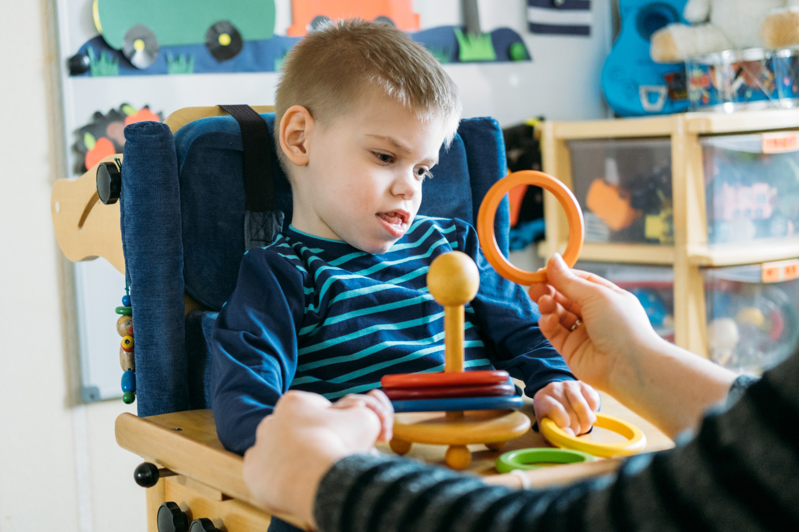 A child in a high chair playing with colorful toys, assisted by an adult hand.