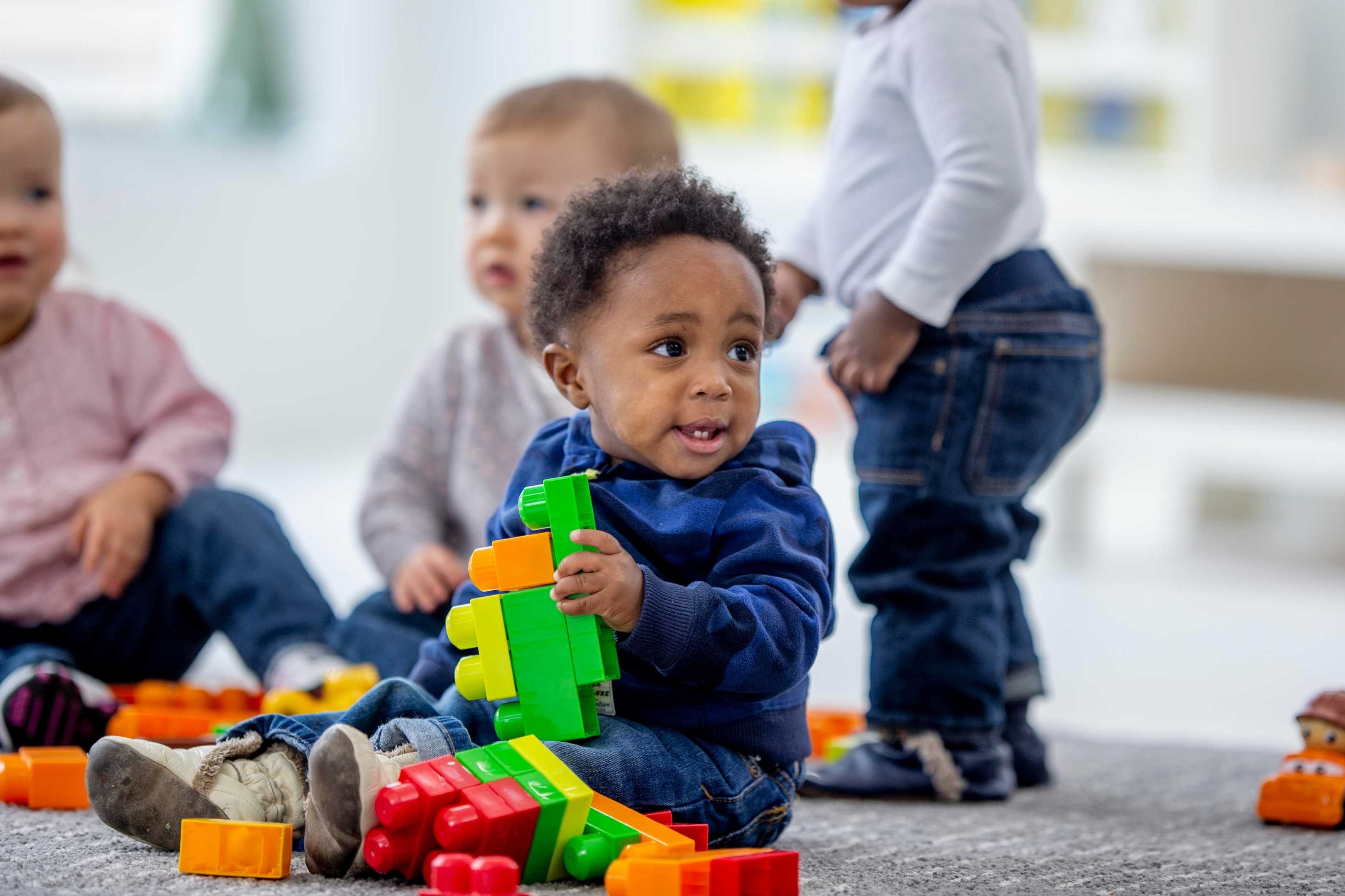 Four children sitting on the floor, playing with toys. One child holds a green toy. Background includes shelves and a wall.