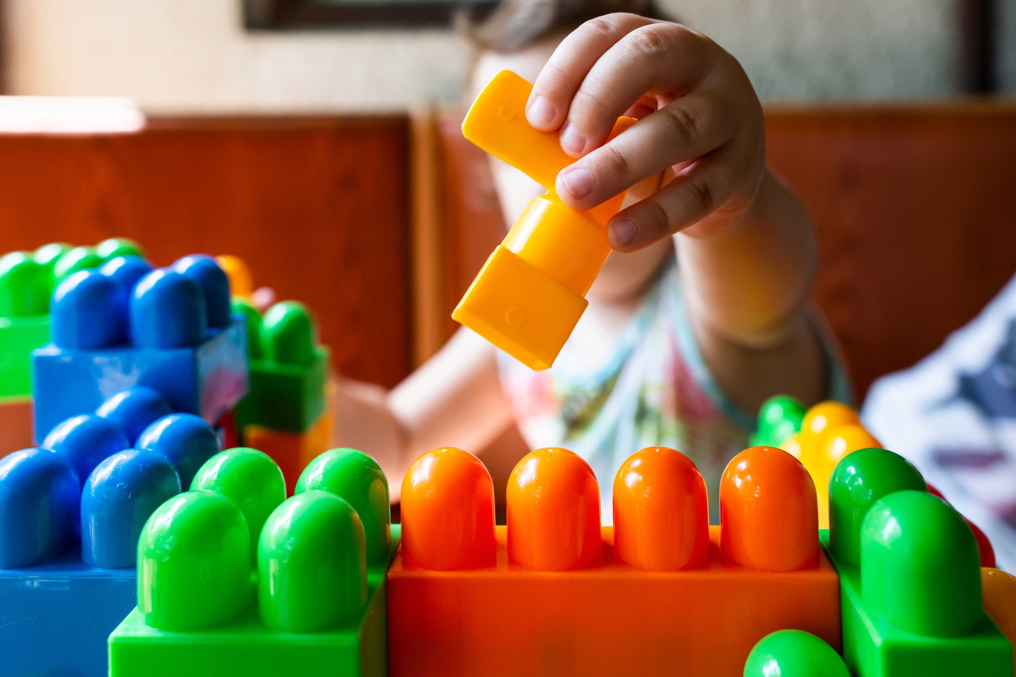 A child plays with colorful building blocks in a room with wooden bench seating in the background.