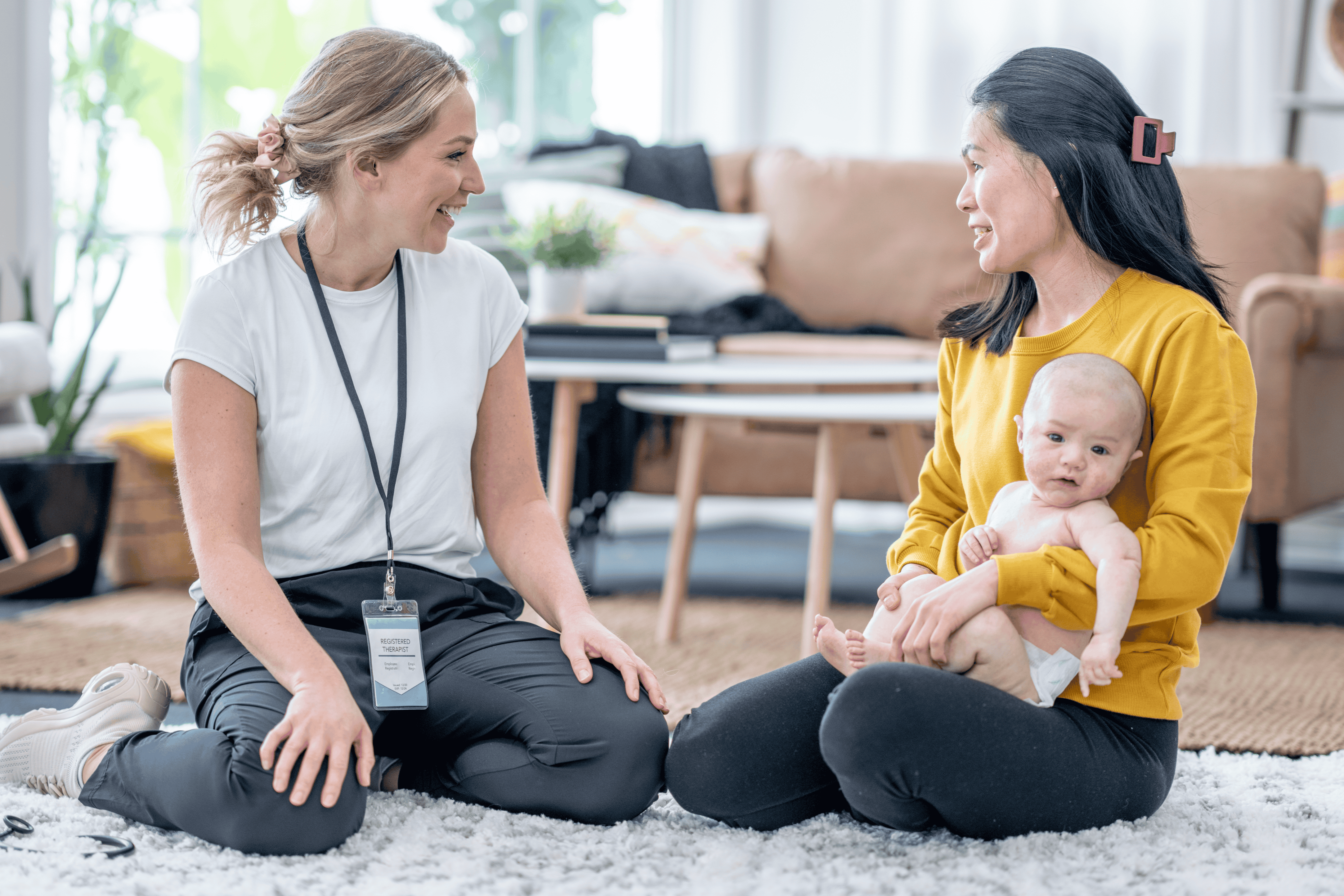 Two women sit on a carpeted floor; one holds a baby. The room has a couch, coffee table, and plants.
