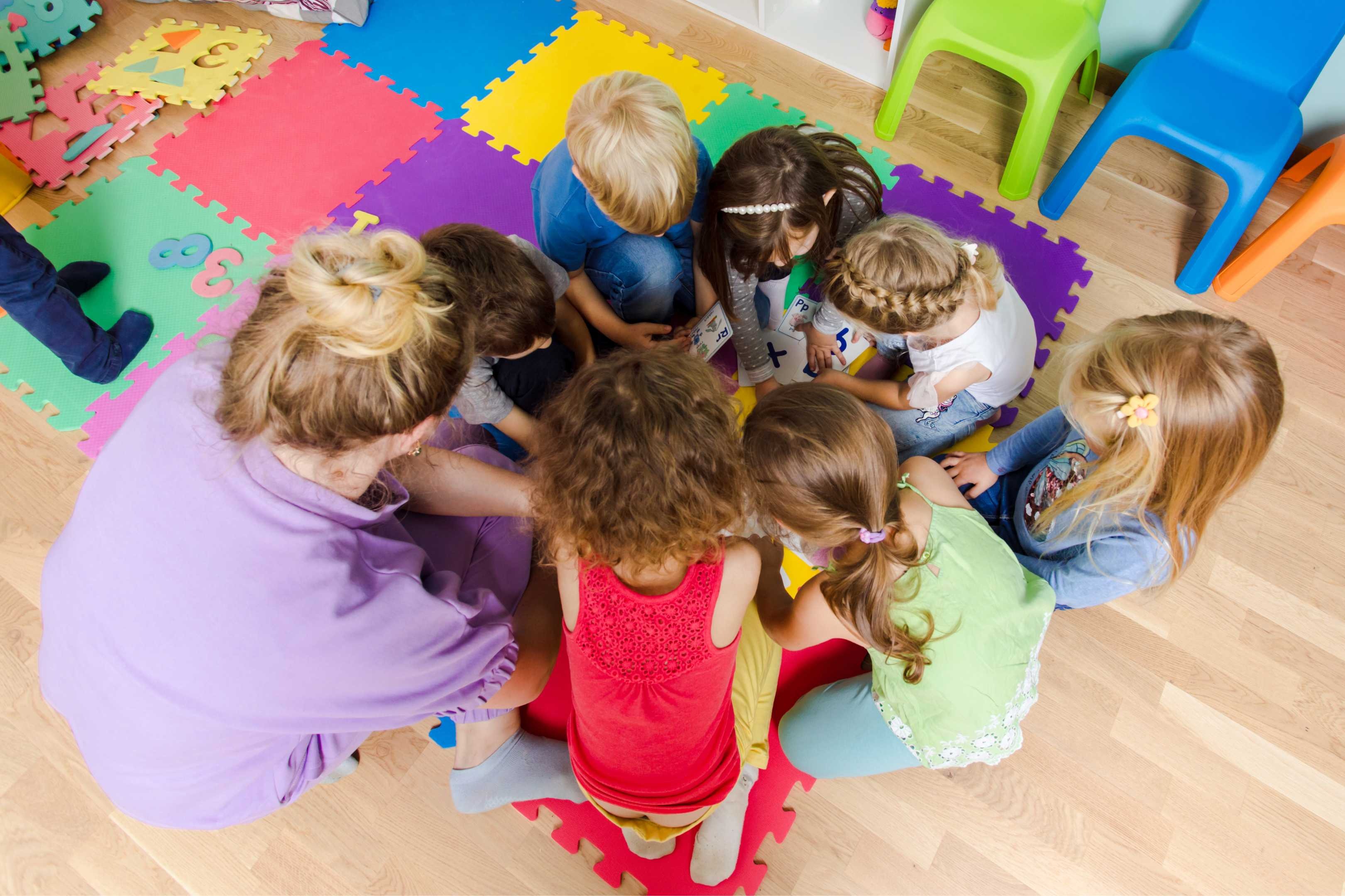 A group of children and two adults sitting in a circle on a wood floor. Colorful chairs and mats are in the background.