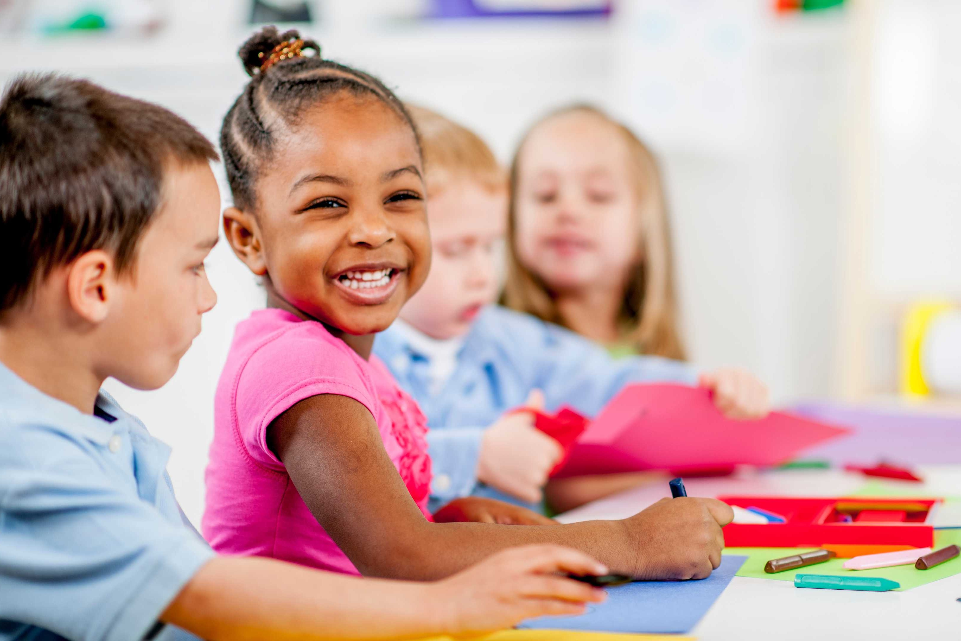 Four children at a table, smiling and holding colorful papers and scissors. A bright room with white walls.