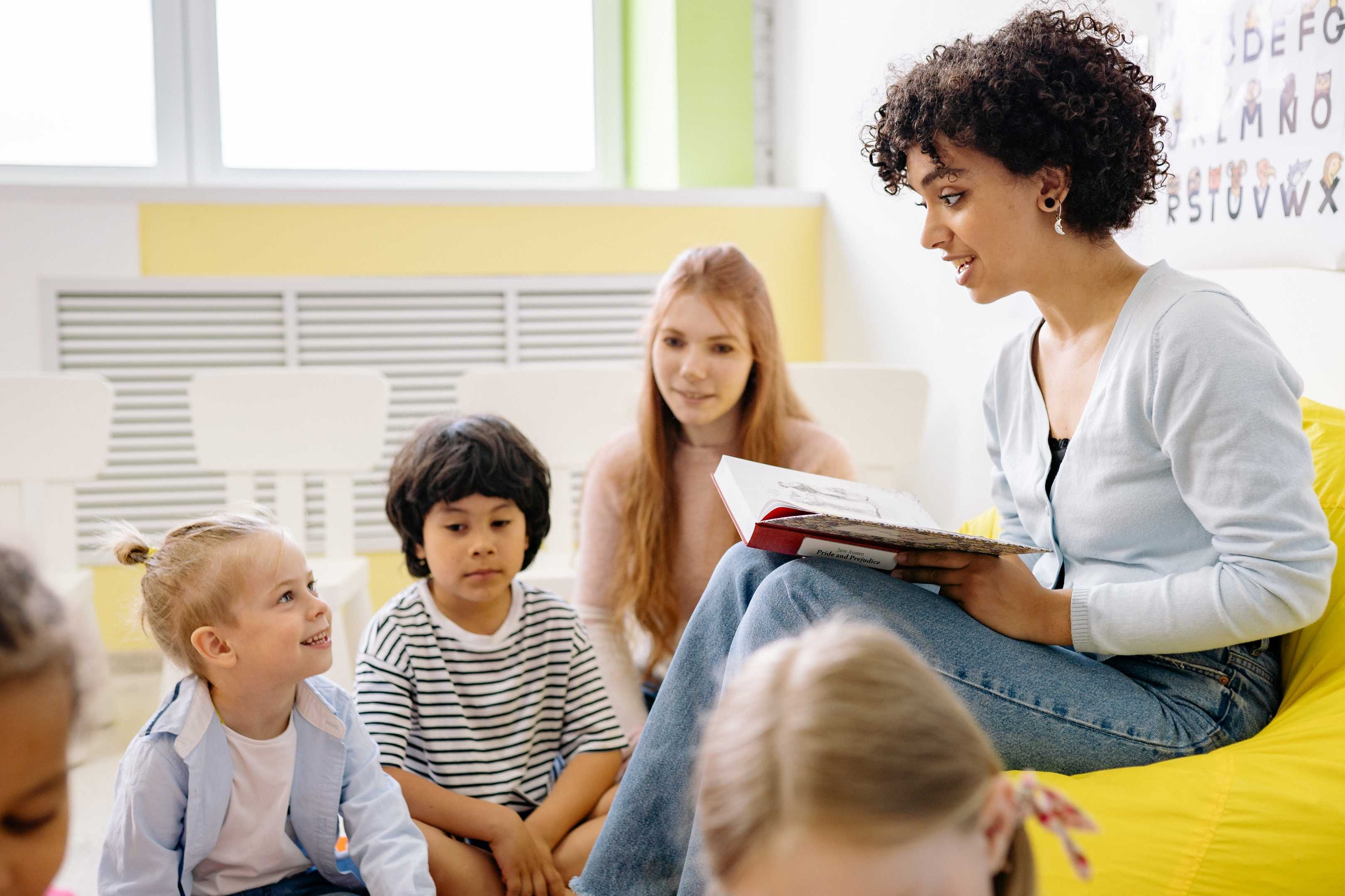 A woman reads a book to a group of children in a classroom with white walls and yellow beanbag chairs.
