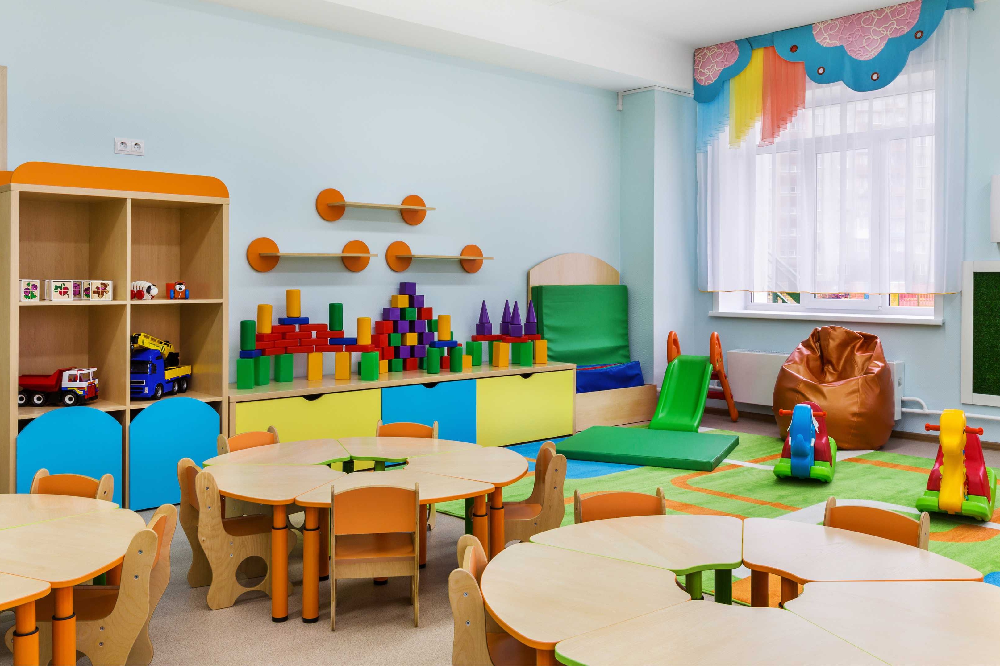 Brightly colored playroom with tables, chairs, shelves, toys, a carpeted area, and a window with blinds.