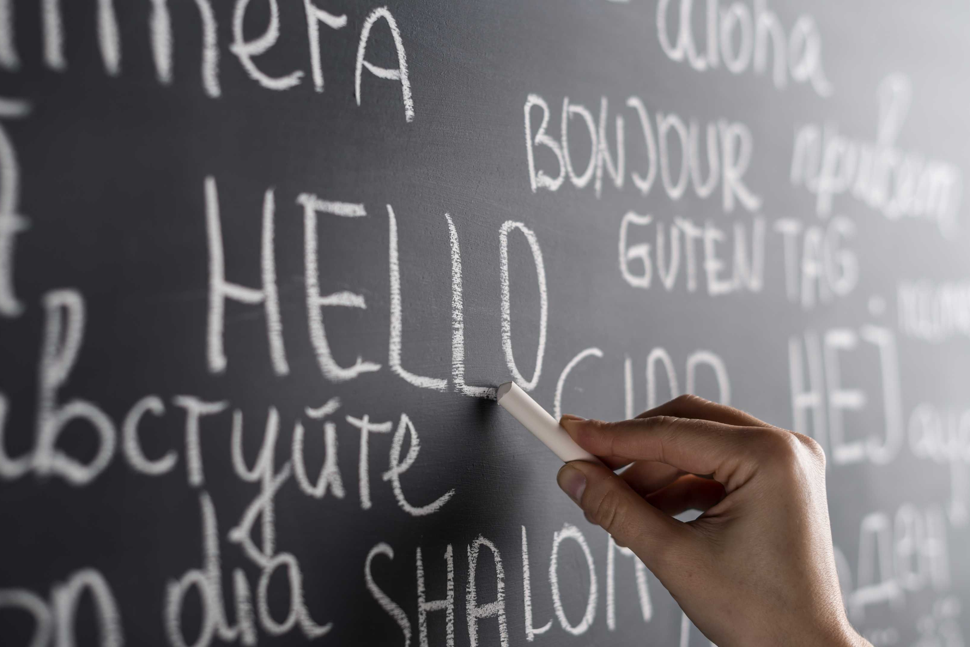A hand writes on a blackboard with various words in different languages, including "HELLO" and "BONJOUR."