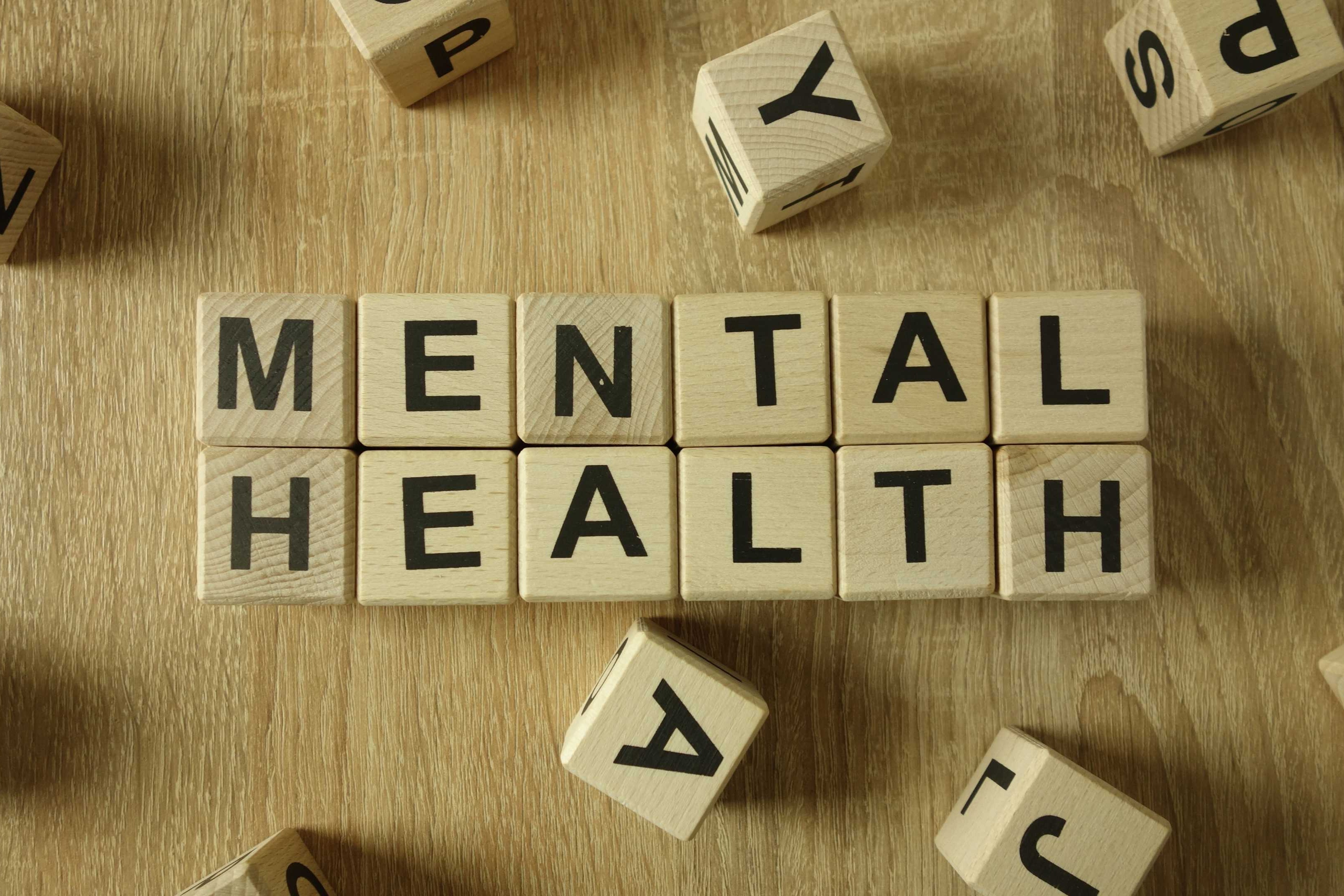 Cubes with letters spelling "MENTAL HEALTH" on a wooden surface, with other letters scattered around.