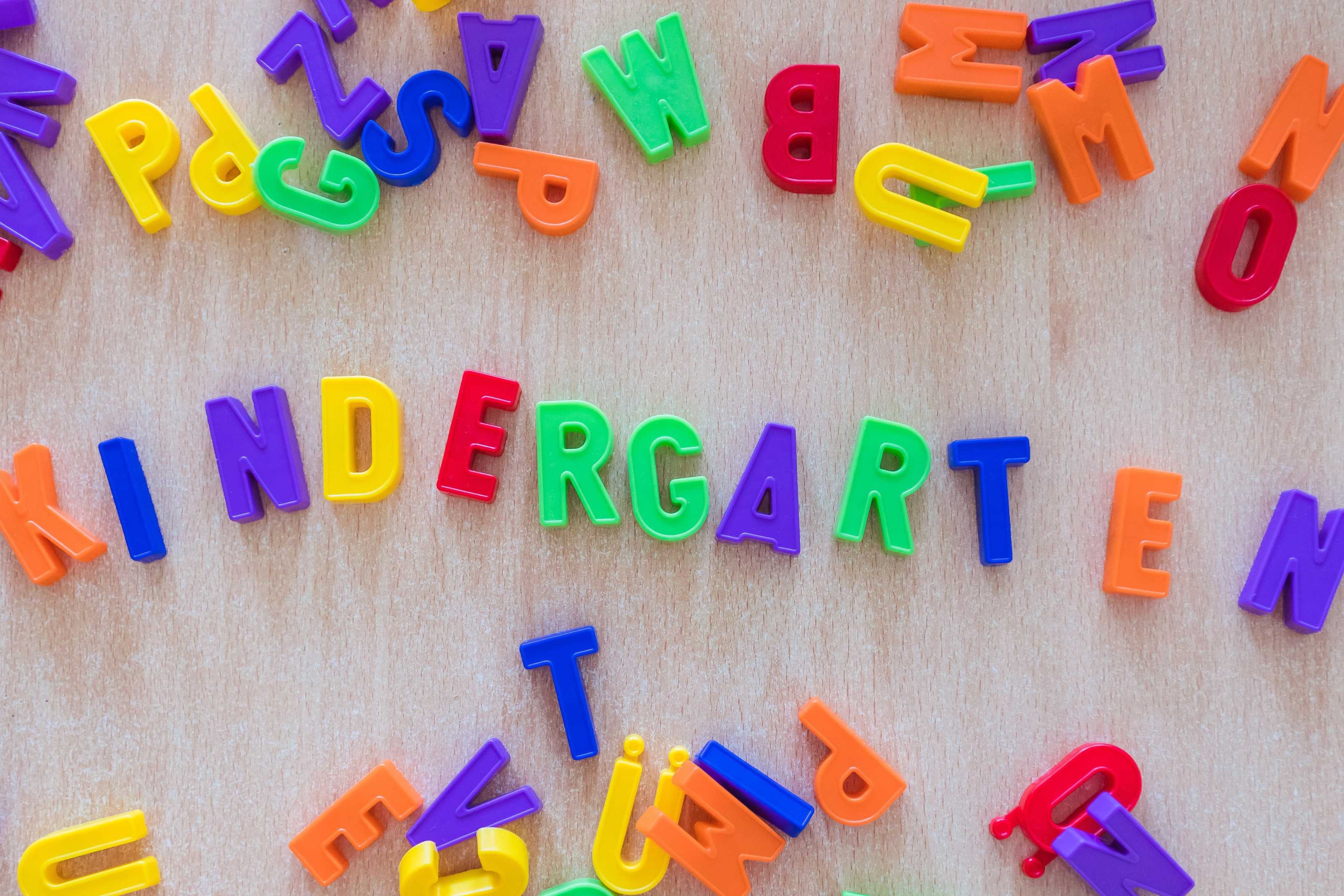 Brightly colored letters on a wooden surface, arranged to spell "Kindergarten" with scattered other alphabet pieces.
