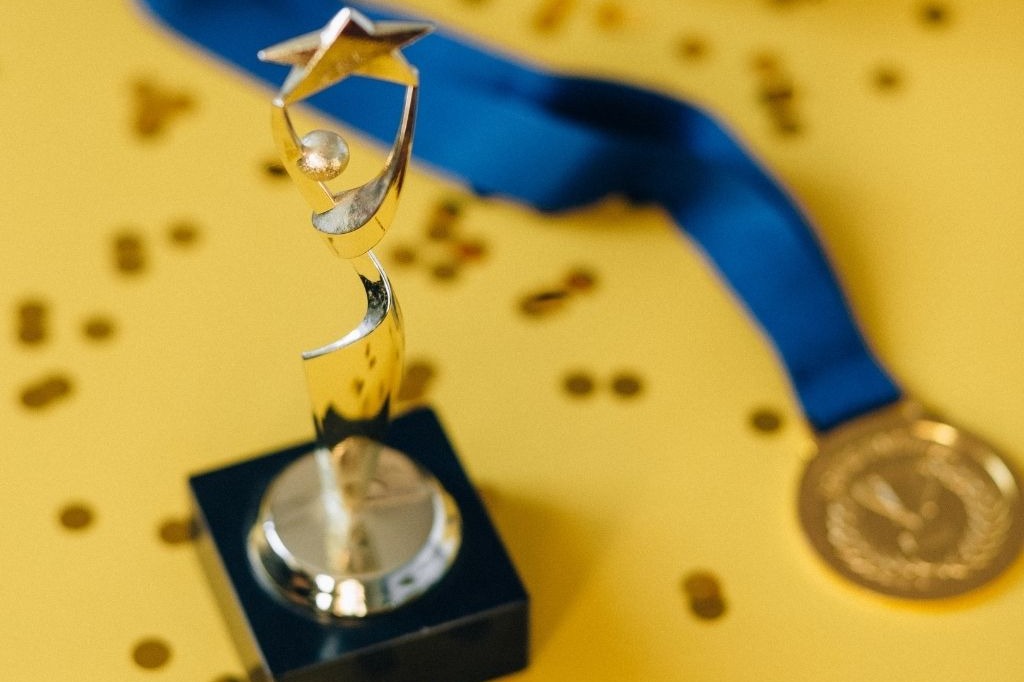 A shiny trophy with a star on top, blue ribbon, and a gold medal lie on a yellow surface with confetti.