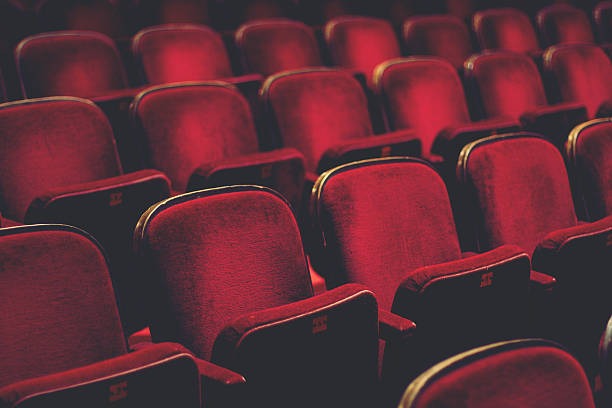 A theater with red velvet seats in rows. The seats are empty, and the theater is dark.