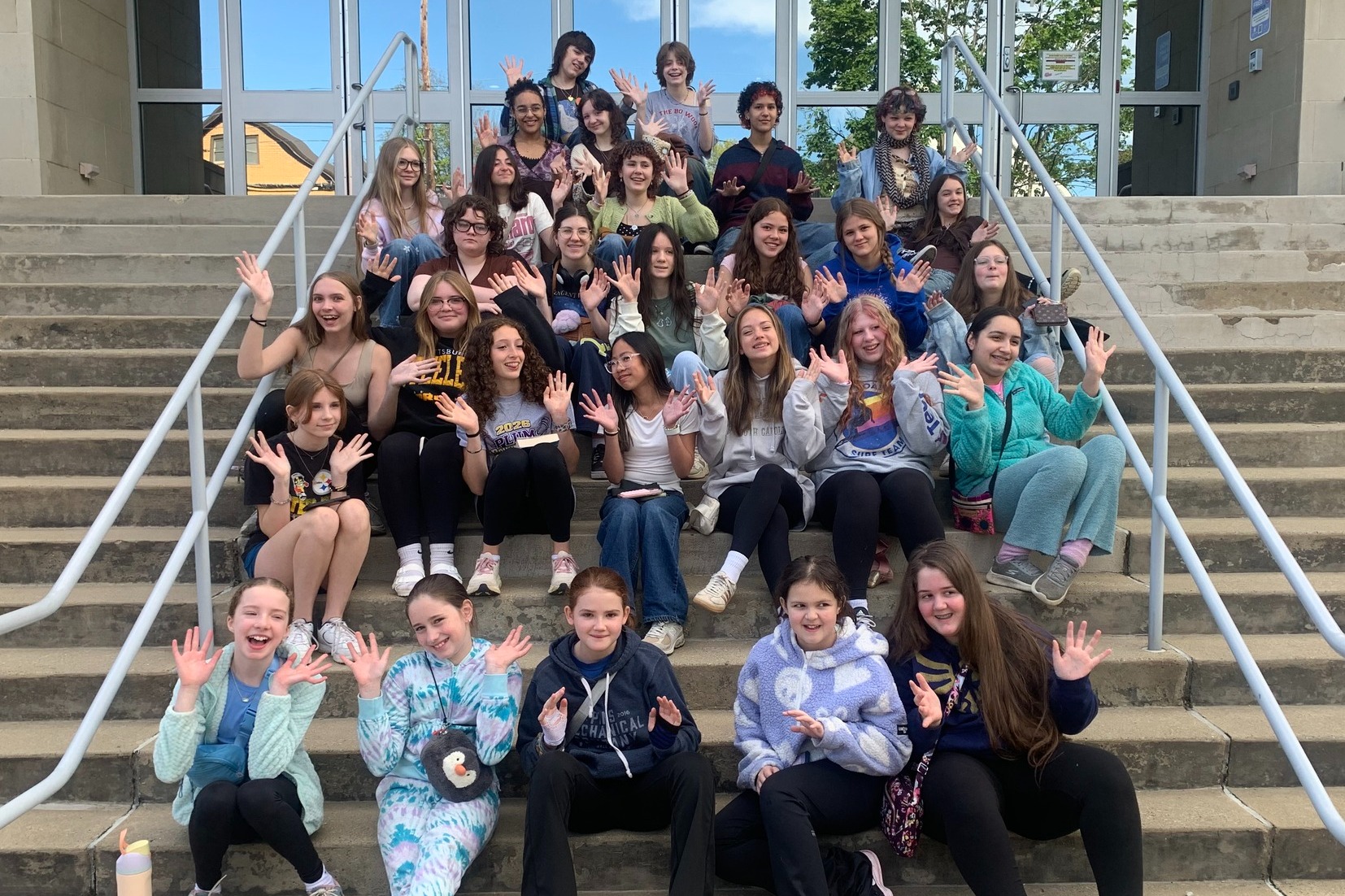 Group of people wearing casual clothes, seated and standing on concrete stairs, smiling for a photo.