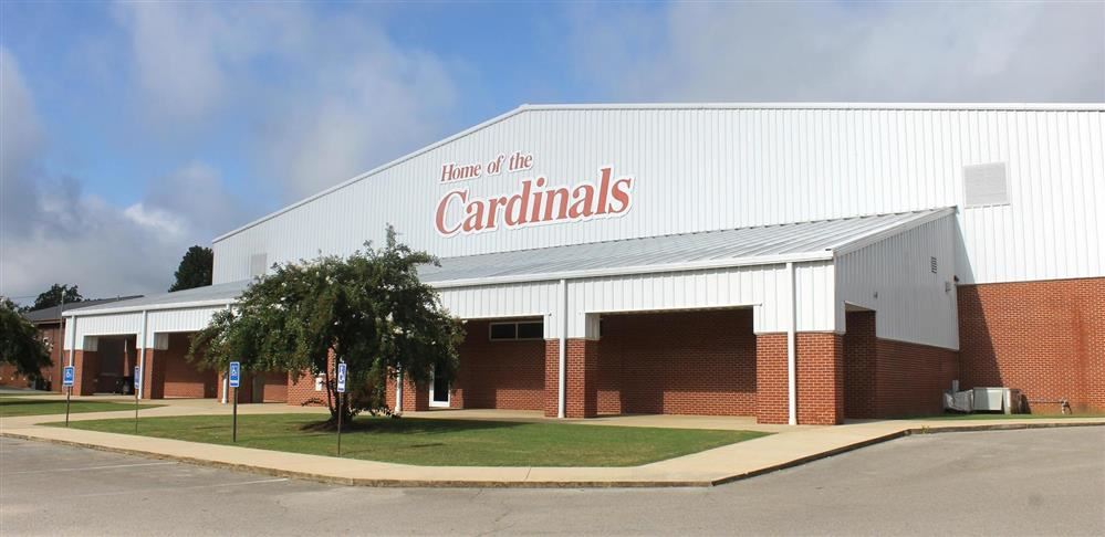The exterior of a large school gymnasium building with red brick on the lower half and white metal siding on the upper half. The words "Home of the Cardinals" are written in red script across the upper front facade.