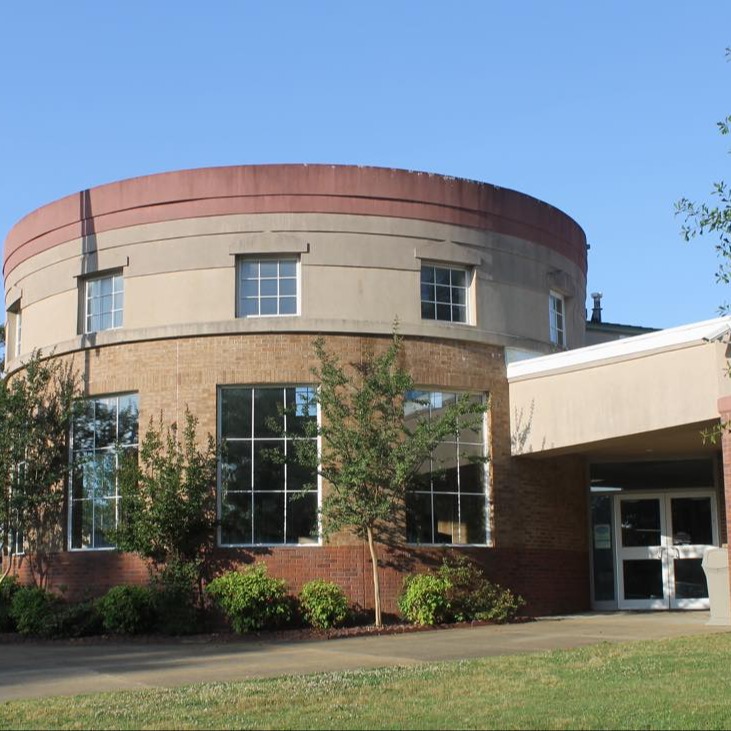 The exterior of a brick building featuring a prominent two-story cylindrical structure with large grid windows on the ground level and smaller square windows on the concrete upper tier. An American flag stands on a tall flagpole to the left, and a covered entrance is visible on the right.