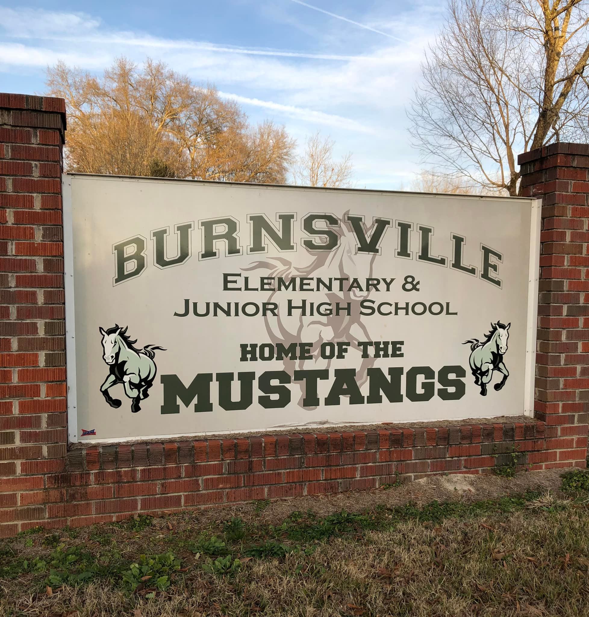 A large outdoor school sign framed by red brick pillars that reads, "Burnsville Elementary &amp; Junior High School, Home of the Mustangs" in green text, flanked by illustrations of two running horses.