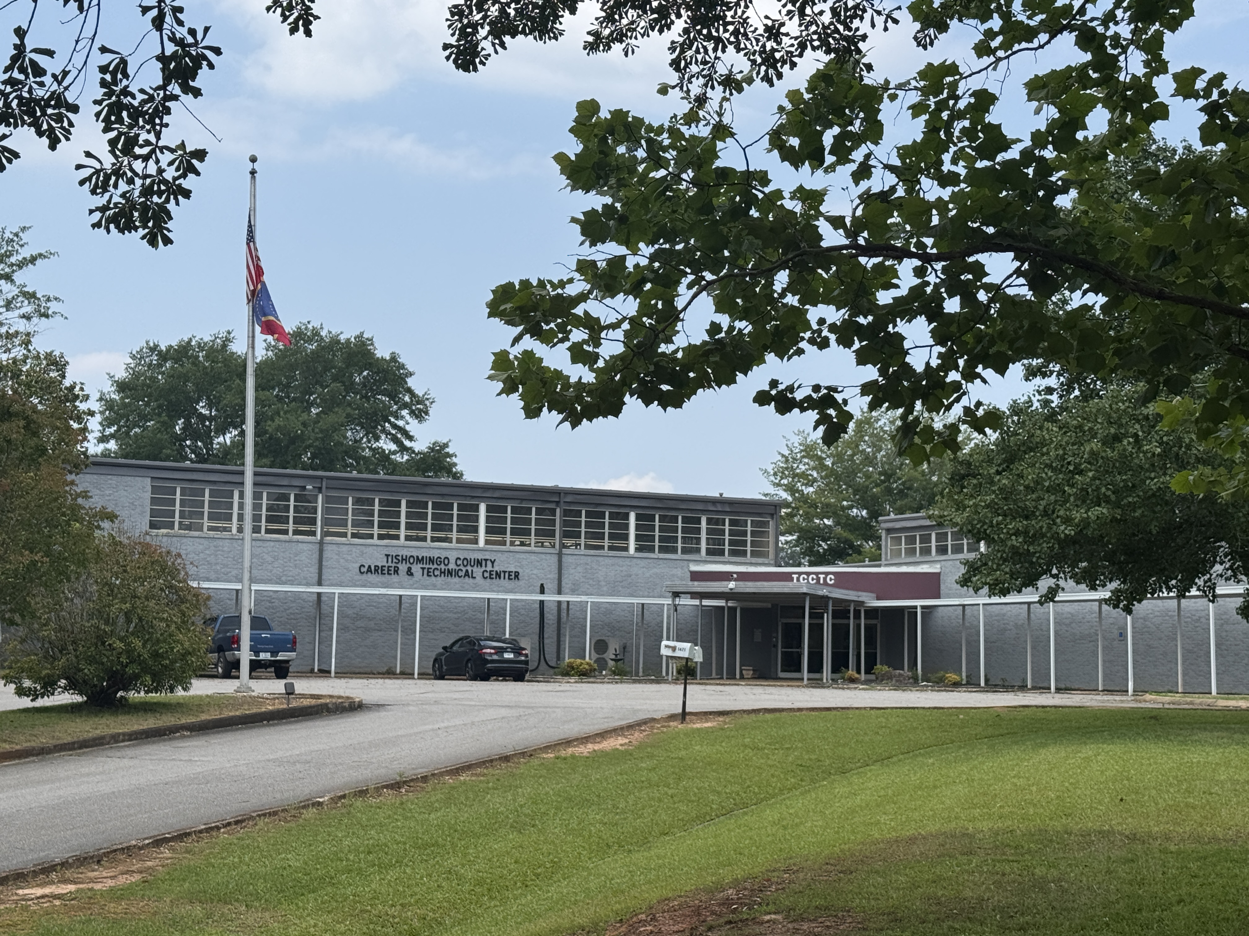 The exterior of the Tishomingo County Career & Technical Center building, a long two-story gray structure with a covered front walkway, a flagpole on the left, and a grassy lawn in the foreground framed by tree branches.