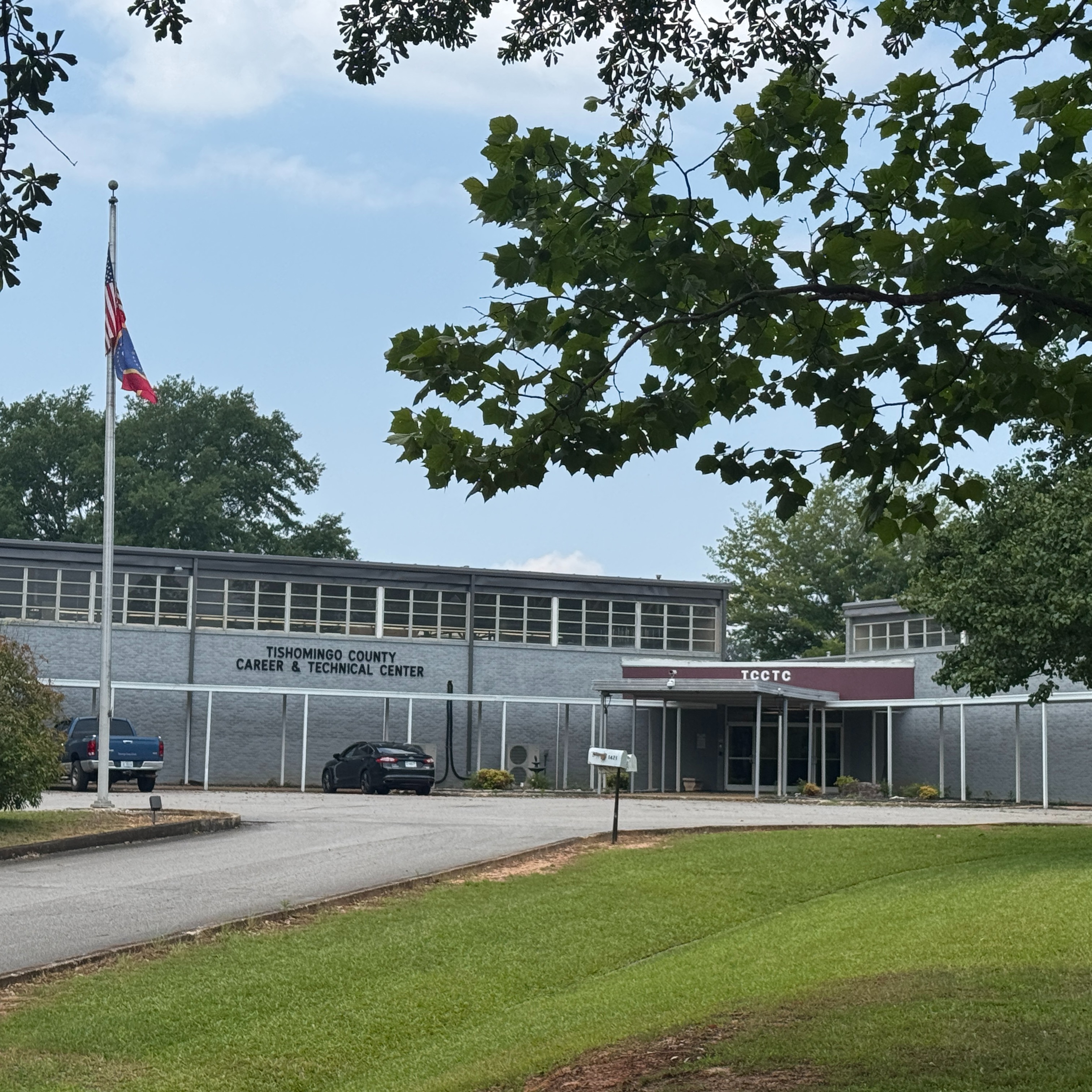 The exterior of the Tishomingo County Career & Technical Center building, a long two-story gray structure with a covered front walkway, a flagpole on the left, and a grassy lawn in the foreground framed by tree branches.