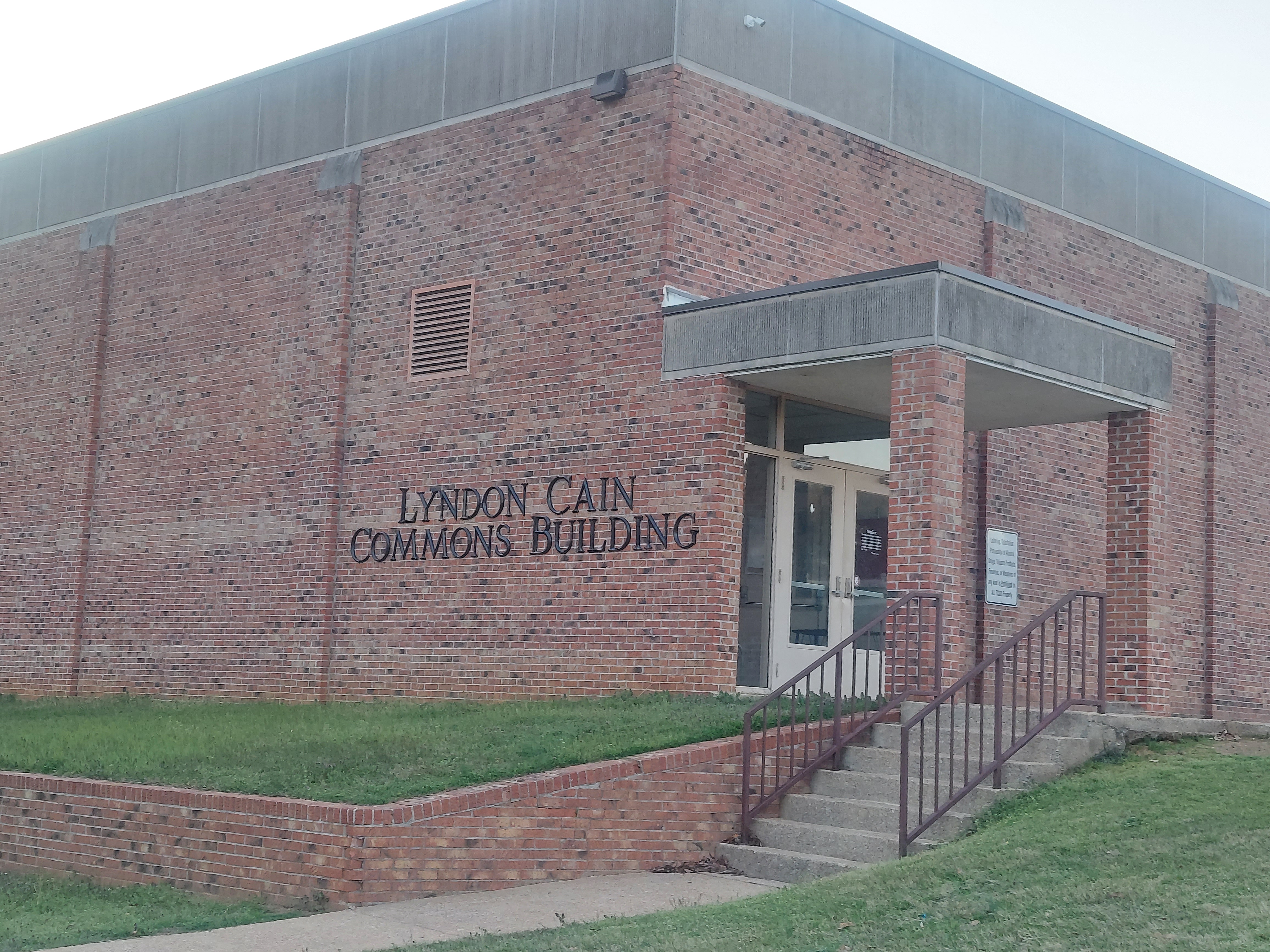The exterior brick entrance of the Lyndon Cain Commons Building, featuring a small flight of concrete stairs with brown metal railings leading up to a covered entryway.