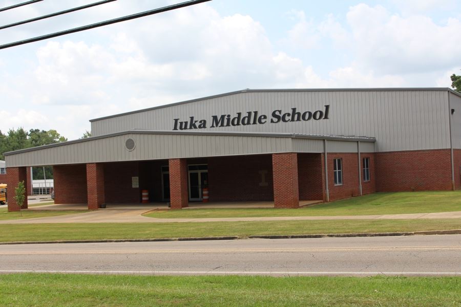 The exterior entrance of Iuka Middle School, featuring a large gym-like building with red brick pillars supporting a covered walkway and light gray metal siding displaying the school's name.