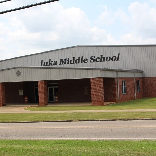 The exterior entrance of Iuka Middle School, featuring a large gym-like building with red brick pillars supporting a covered walkway and light gray metal siding displaying the school's name.